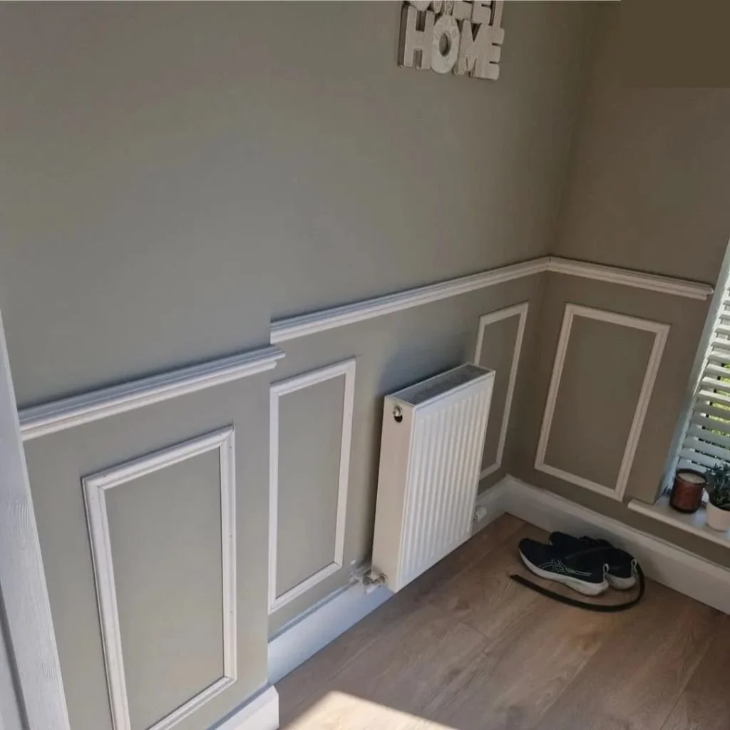 Corner of a room with green walls, white trim, a radiator, a pair of black sneakers with white soles on the floor, and a window with white blinds.