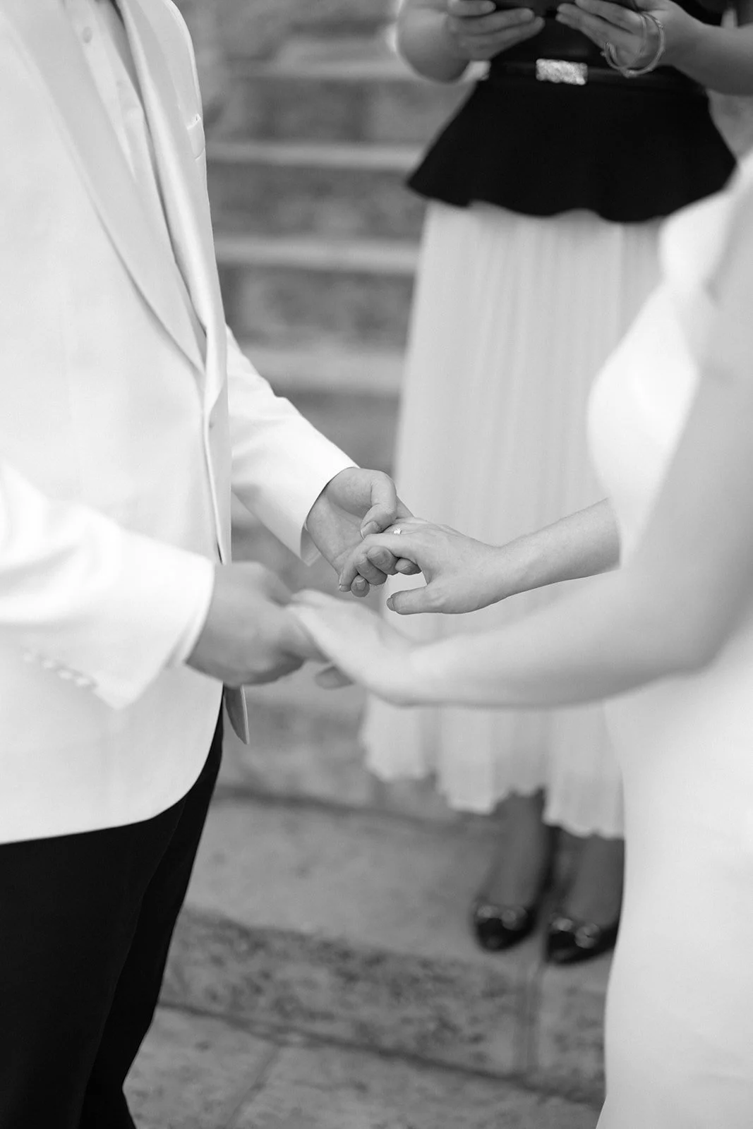 A black and white photo of a couple holding hands during a wedding ceremony, with an officiant standing nearby, and a woman in a white dress and black heels visible in the background.