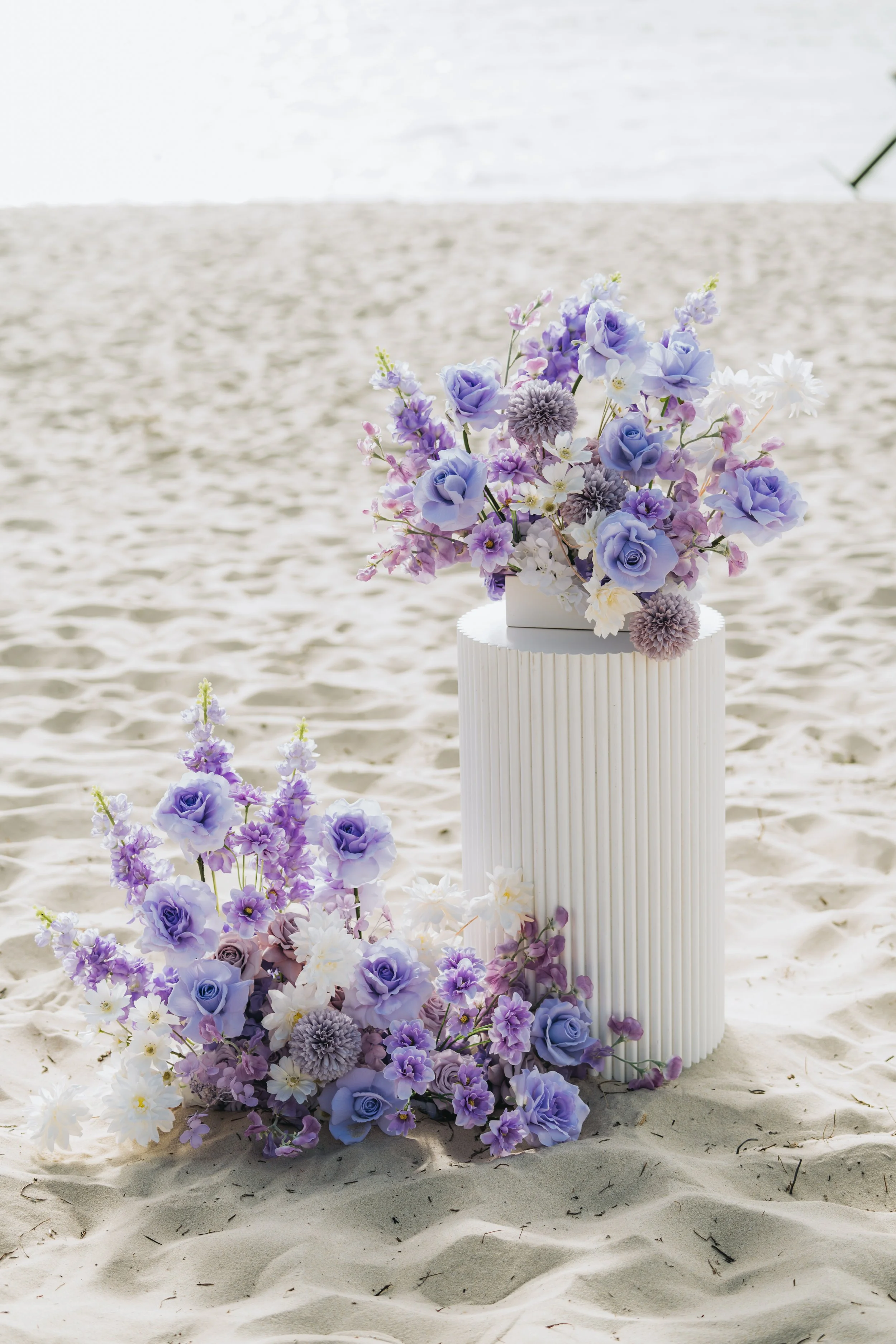 Floral arrangement with lavender, purple, and white flowers on a white pedestal and on the ground on a sandy beach.