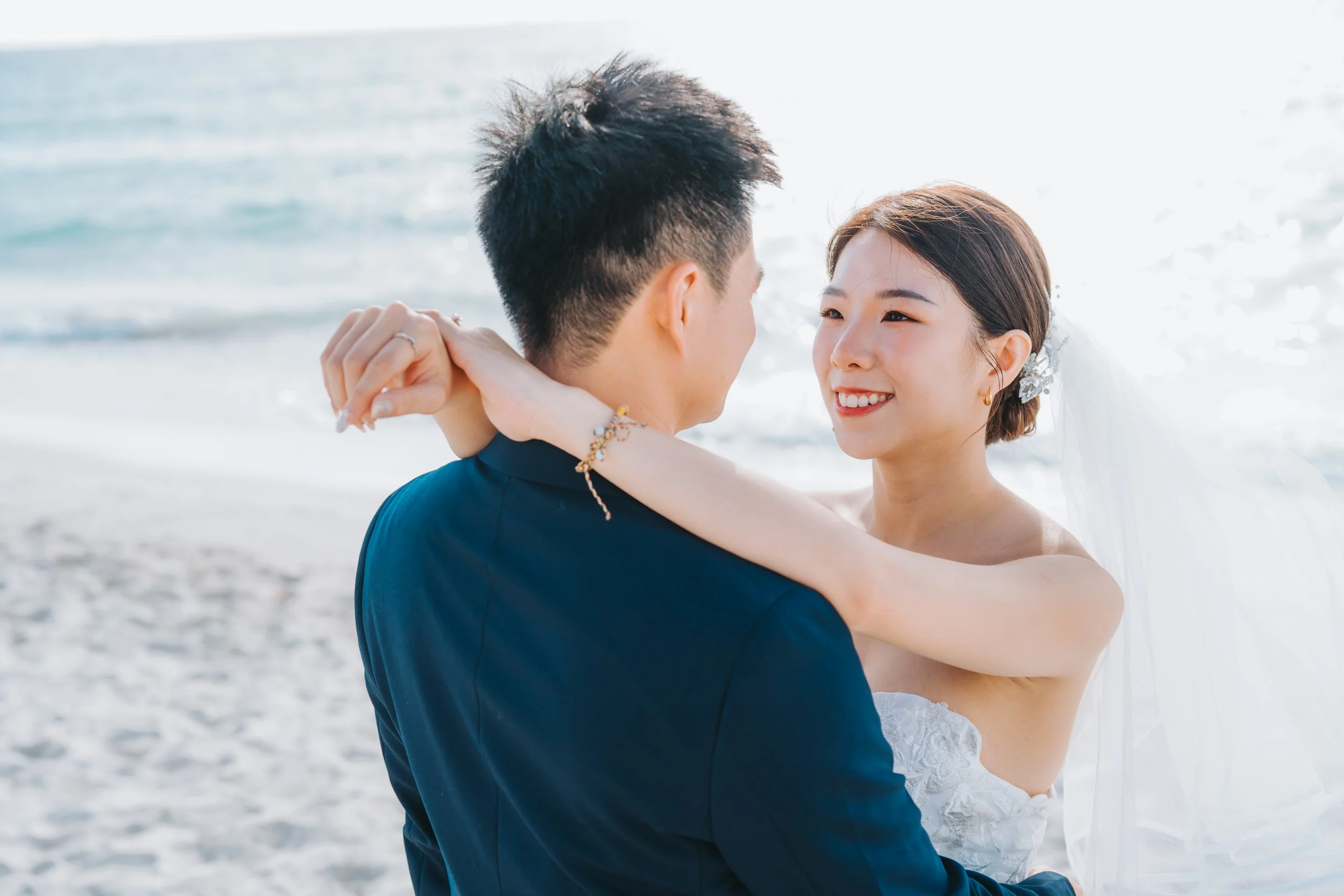 A bride and groom embracing on a beach, gazing into each other's eyes.