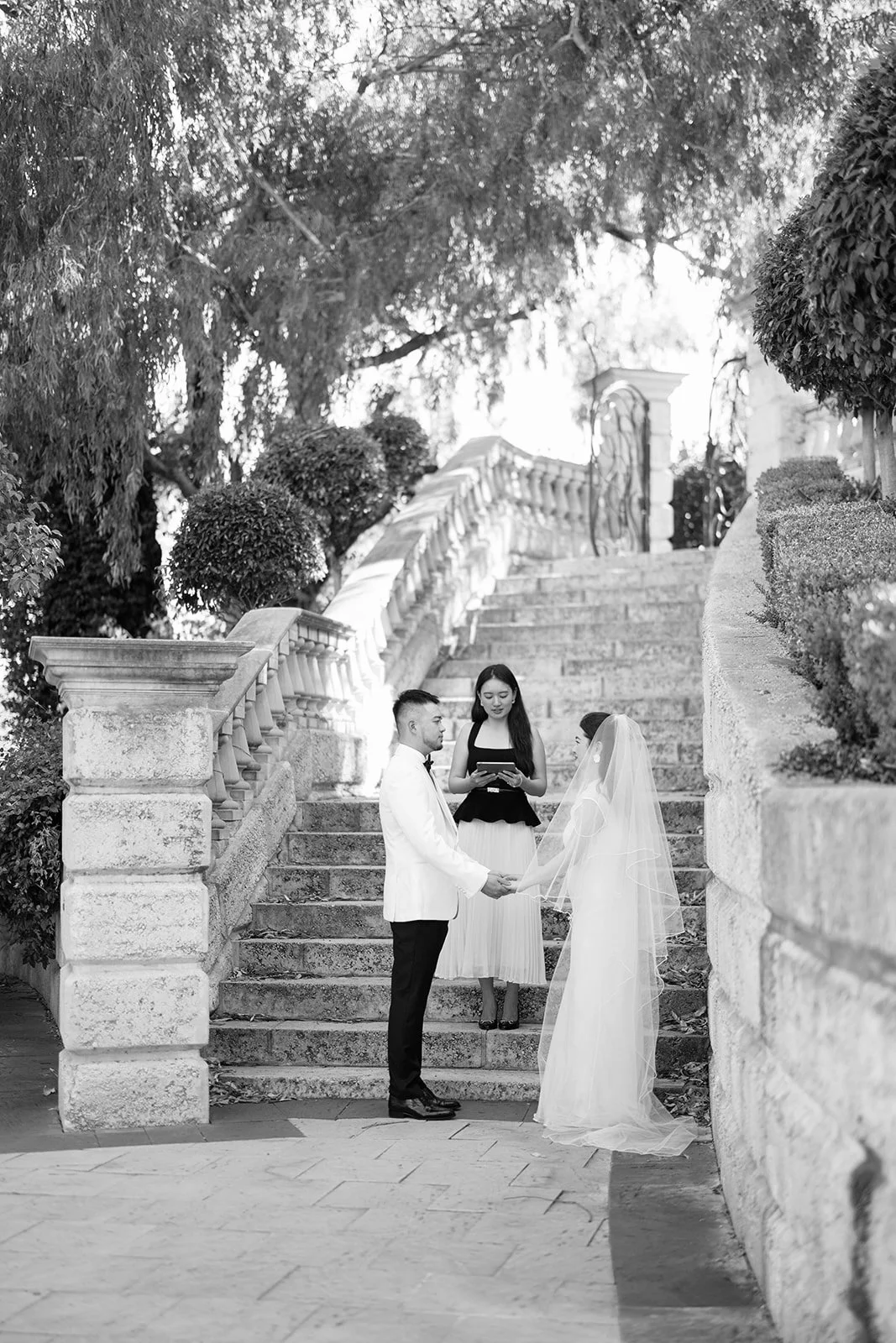 A wedding ceremony taking place outdoors on stone steps, with a bride and groom holding hands and exchanging vows, surrounded by trees and bushes. An officiant stands behind them, overseeing the ceremony.