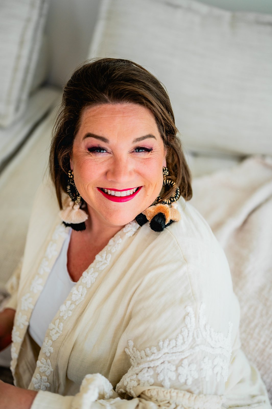 Portret van een lachende vrouw met korte bruine haar, roze make-up, rode lipstick, sieraden en een lichte jasje met geborduurde details, zittend op een lichte sofa.