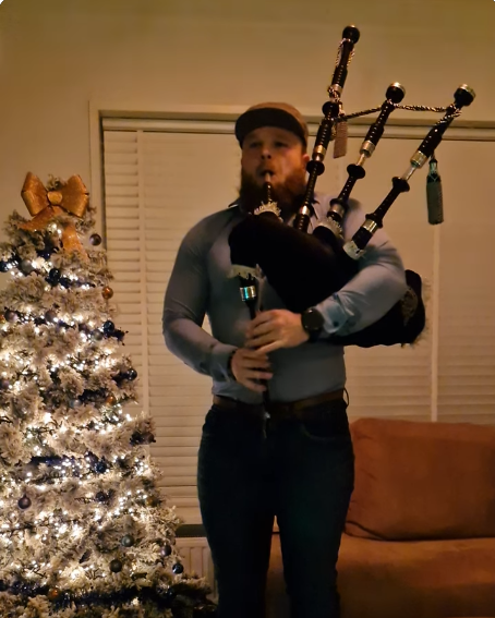 Man die opnamen speelt op een grote doedelzak in een woonkamer naast een versierde kerstboom.