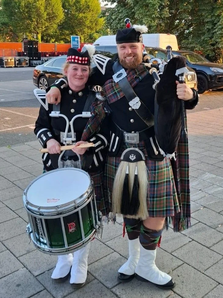 Twee mensen in traditionele Schotse kledij, één met een trommel en de andere met een doedelzak, poseert buiten op straat.