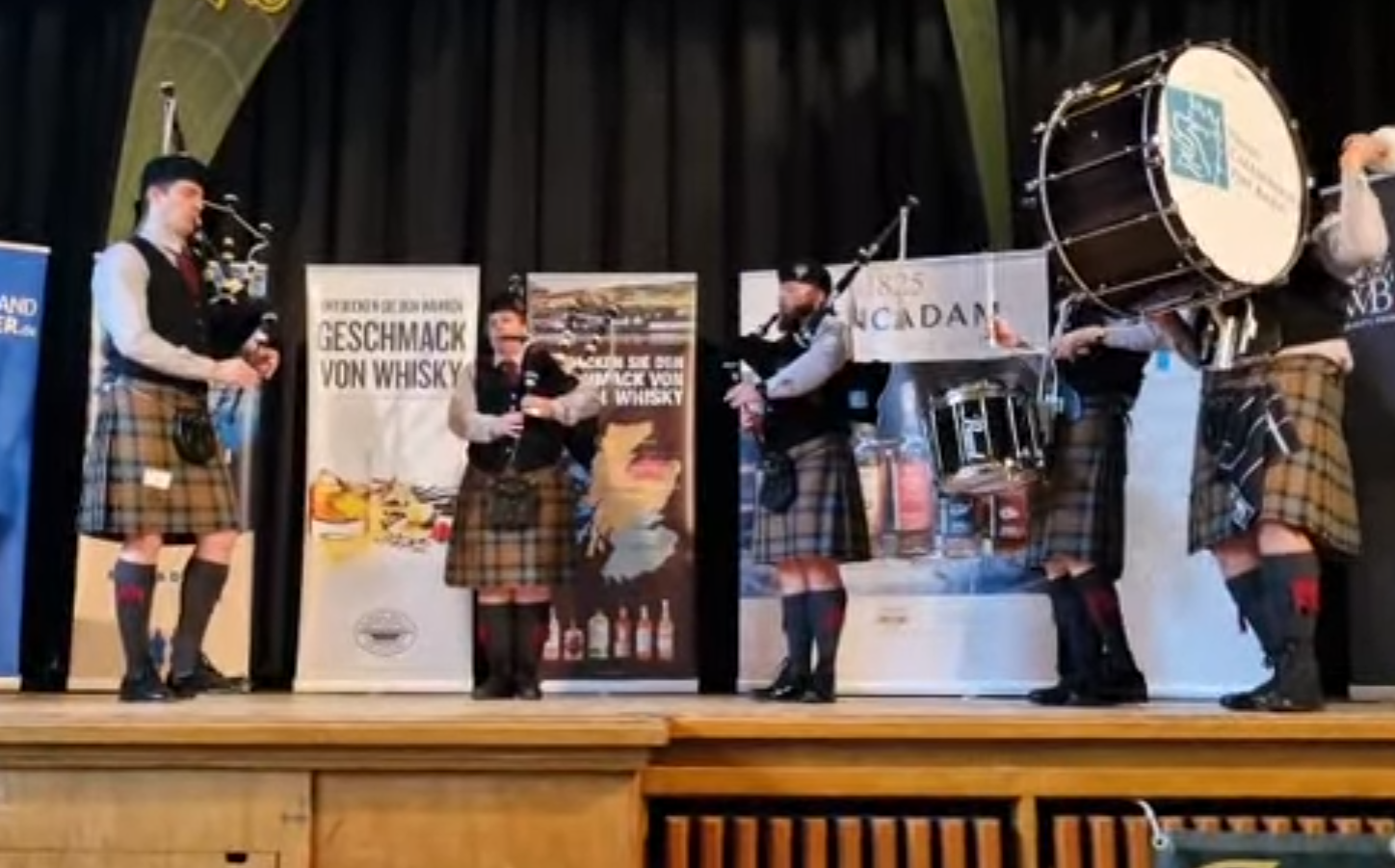 Vijf mannen in traditionele Schotse kilts spelen op muziekinstrumenten op een podium, met banners op de achtergrond.