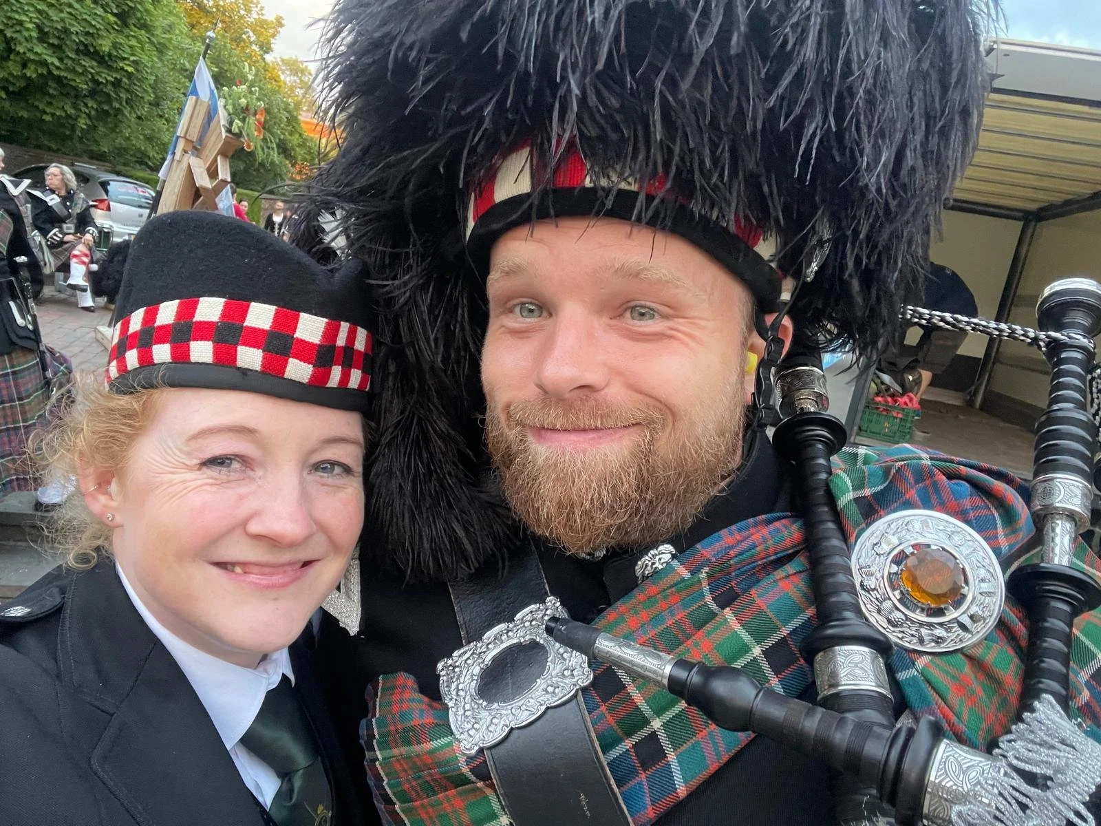 Een vrouw en een man gekleed in traditionele Schotse feestkleding, de man speelt de doedelzak, ze staan dicht bij elkaar en glimlachen. Op de achtergrond zijn mensen en auto's te zien.