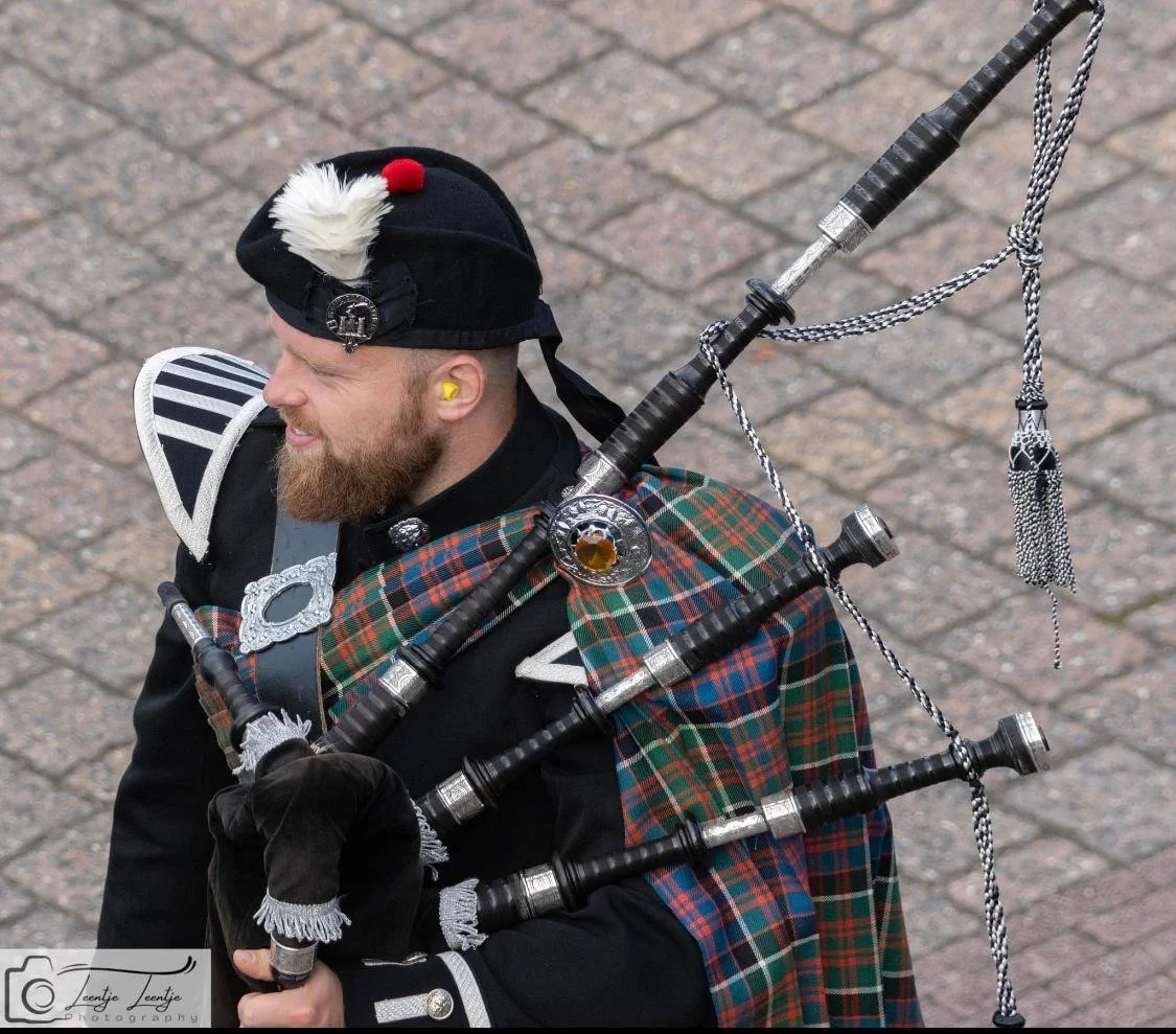 Man in traditionele Schotse kleding speelt de doedelzak, draag een kilt, jas en een historisch uniform, en loopt op een stenen straat.