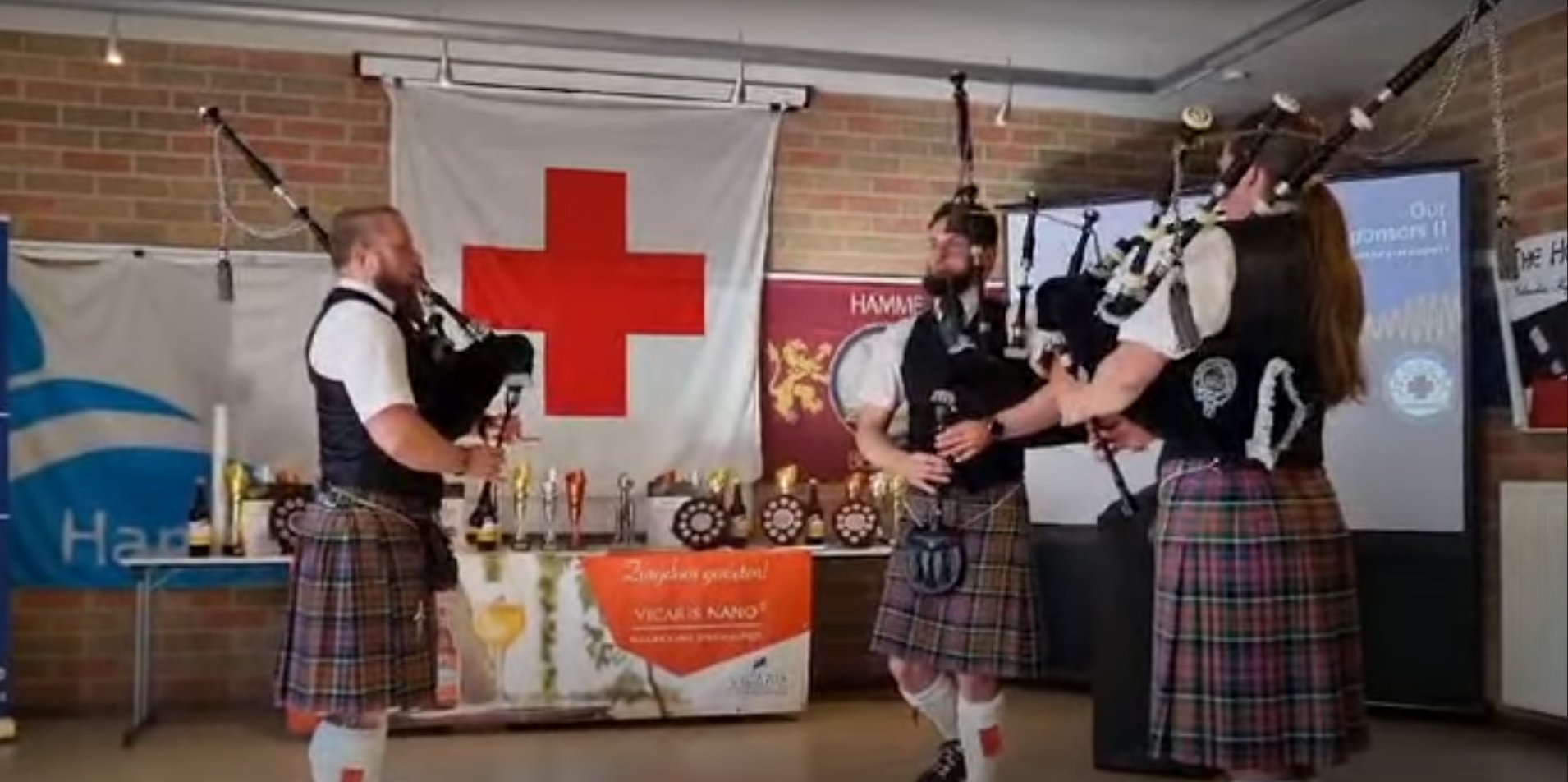 Drie mensen in traditionele Schotse kleding die muzikaal optreden met bagpipes in een zaal met verschillende banners en een tafel met drankjes en prijzen, vlag van de Schotse Clan MacGregor op de achtergrond.