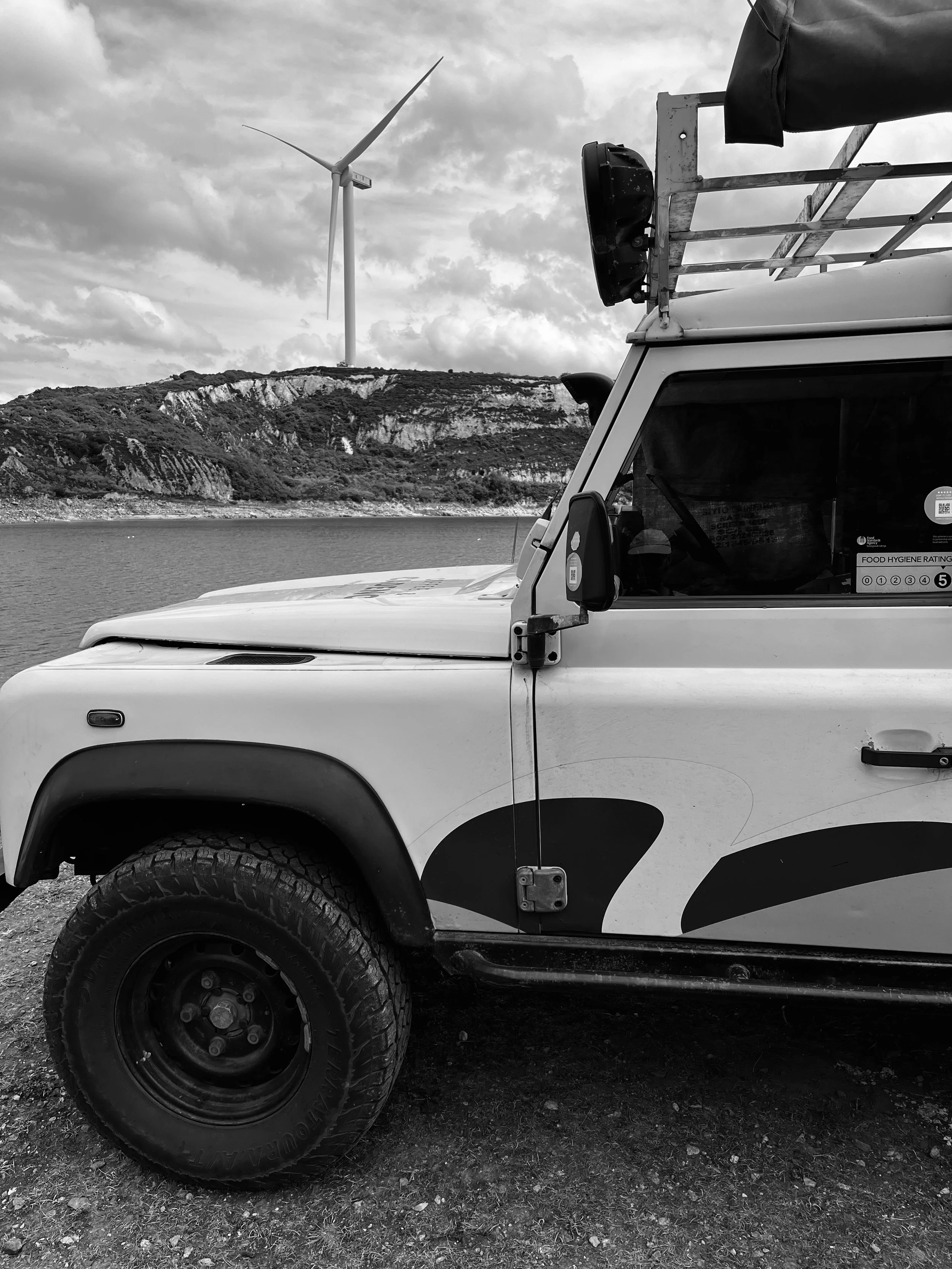 A section of a white off-road vehicle with a spare tire, antenna, and a roof rack parked near a body of water, with hills and a wind turbine in the background under cloudy skies.