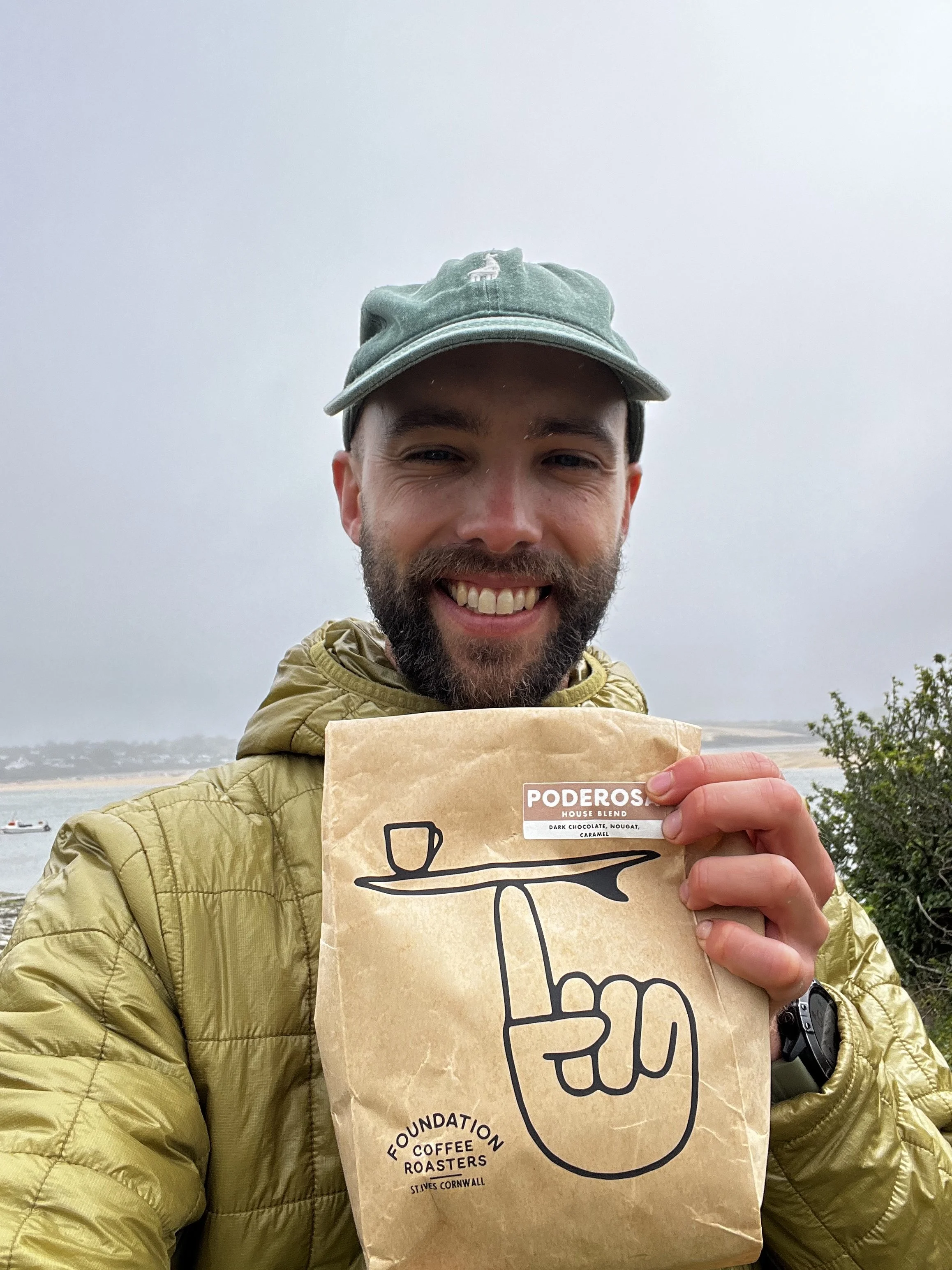 A man with a beard and wearing a green baseball cap and a yellow jacket holding a paper bag with a coffee logo on it, smiling outdoors with a cloudy sky and water in the background.