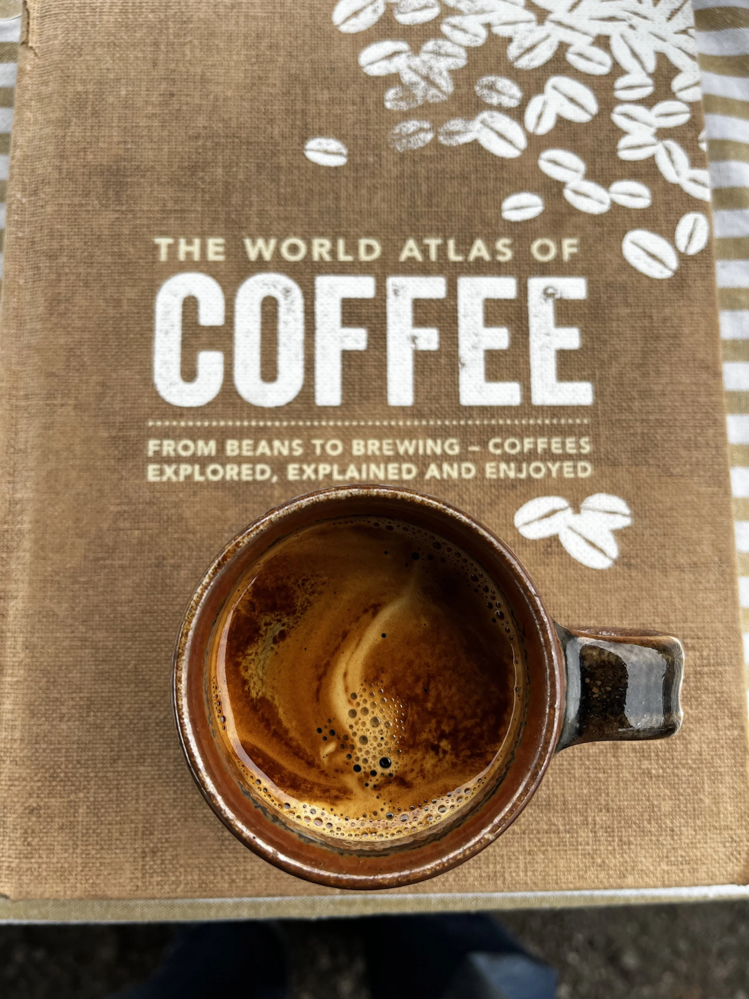 A coffee mug filled with espresso on a book titled 'The World Atlas of Coffee' with coffee beans and a striped cloth in the background.