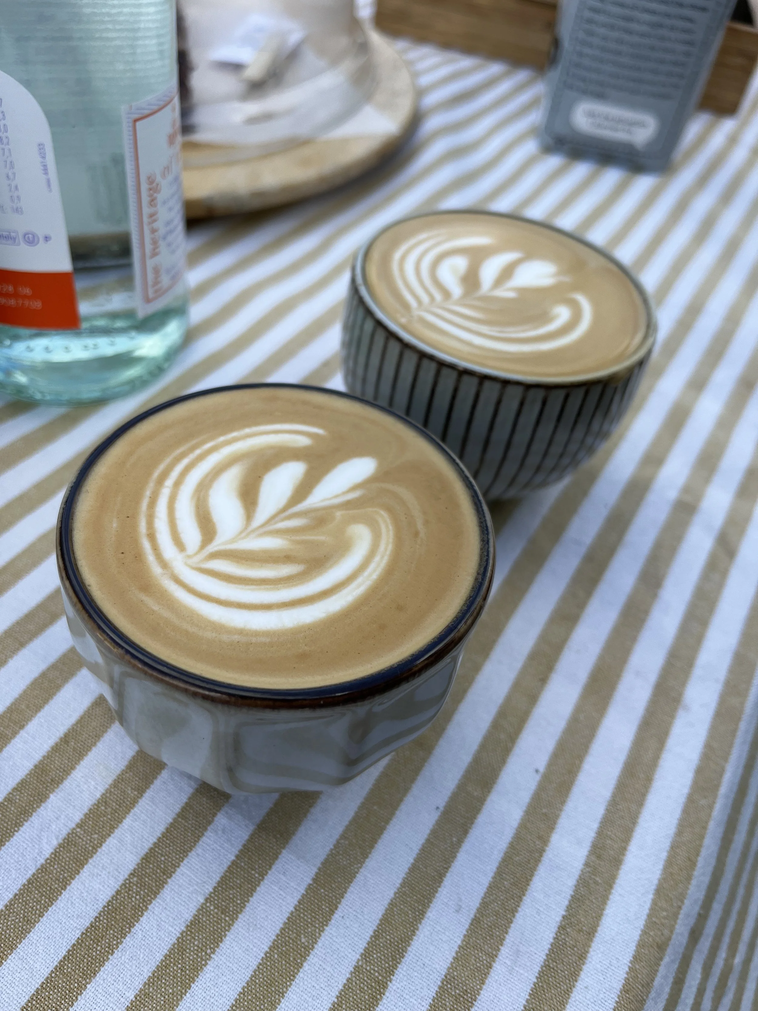 Two cups of coffee with latte art on a striped tablecloth, with a water bottle and some other objects in the background.