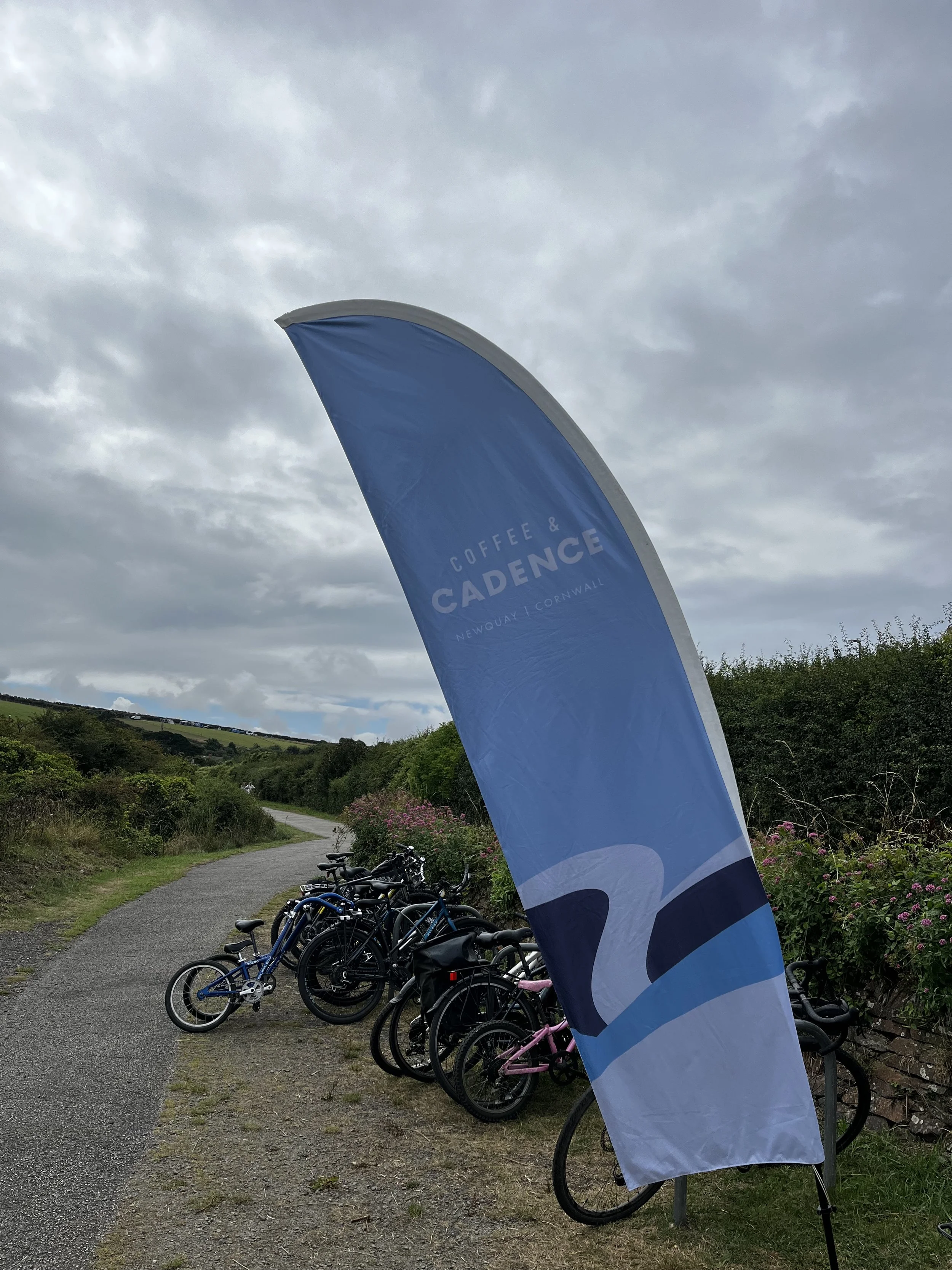 Blue flag with text 'Coffee & Cadence Newquay Cornwall' near a row of parked bicycles on a rural pathway under cloudy sky.