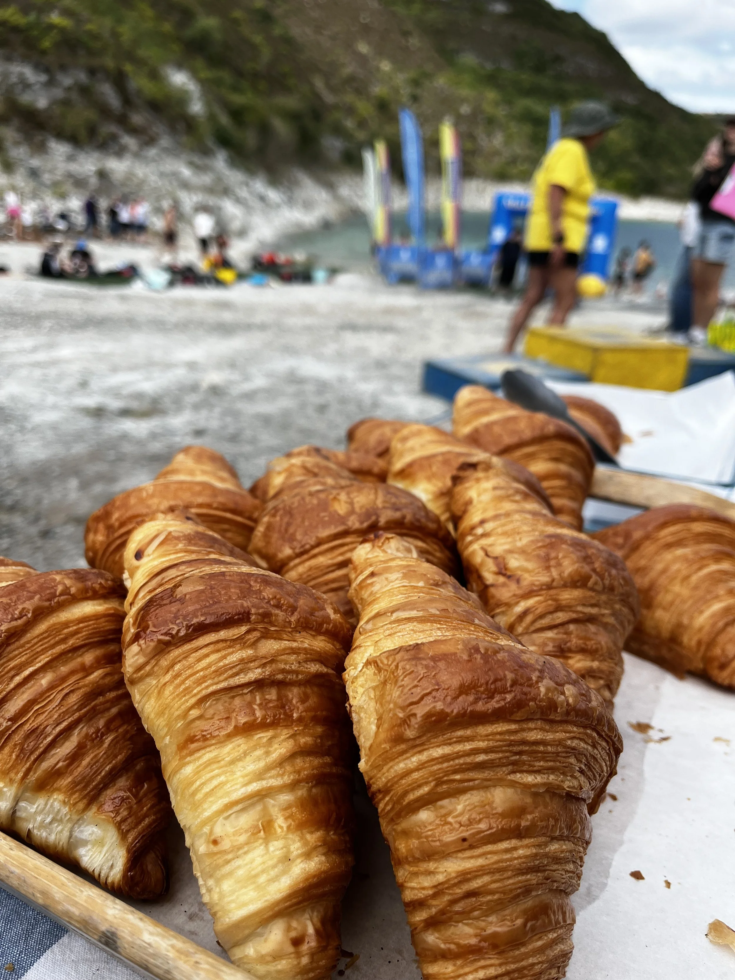Close-up of golden-brown croissants on a tray at the beach with people, colorful flags, and a rocky hill in the background.