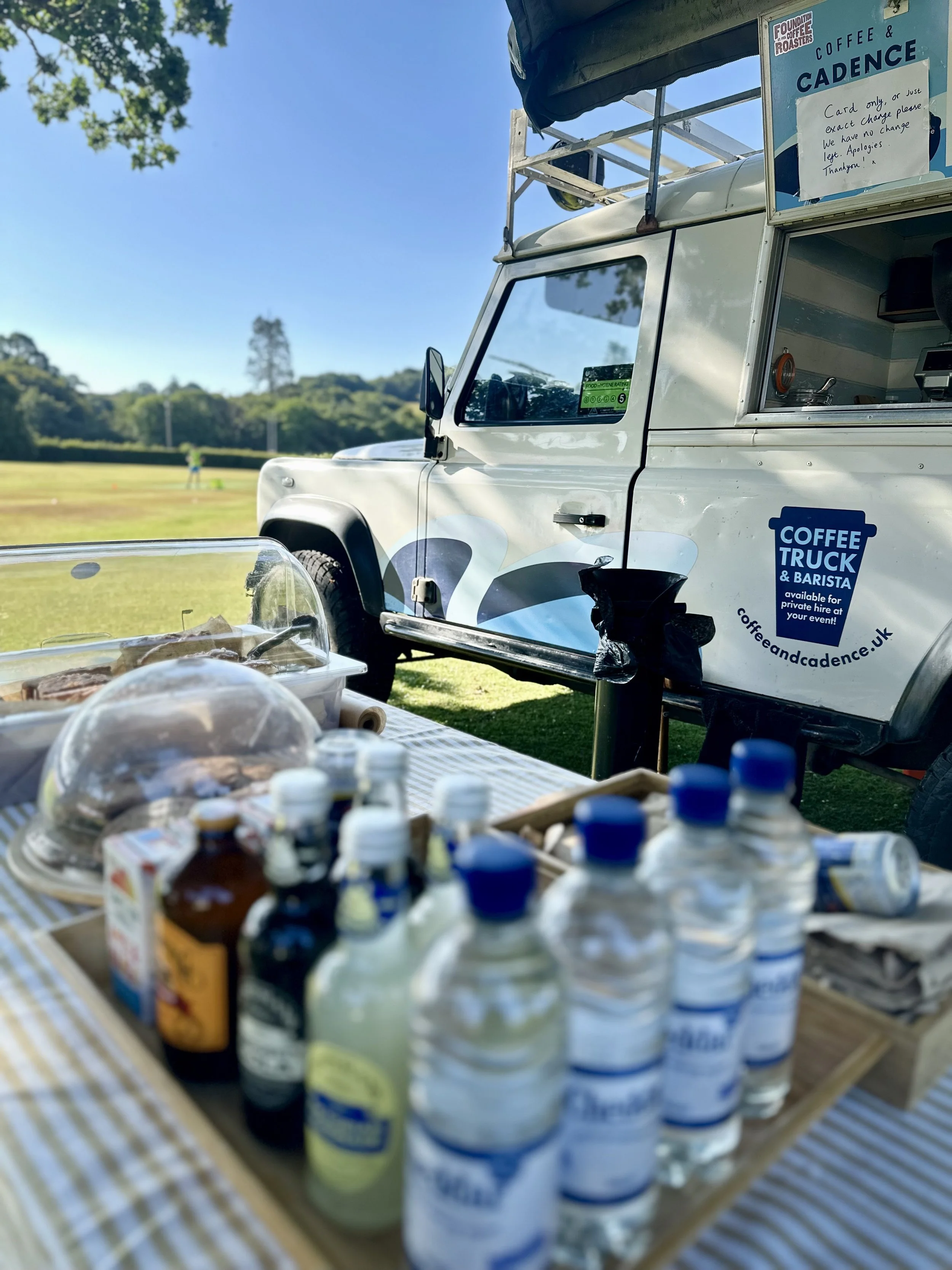 Food and drink supplies on a picnic table next to a coffee truck at an outdoor event, with a grassy field and trees in the background on a sunny day.