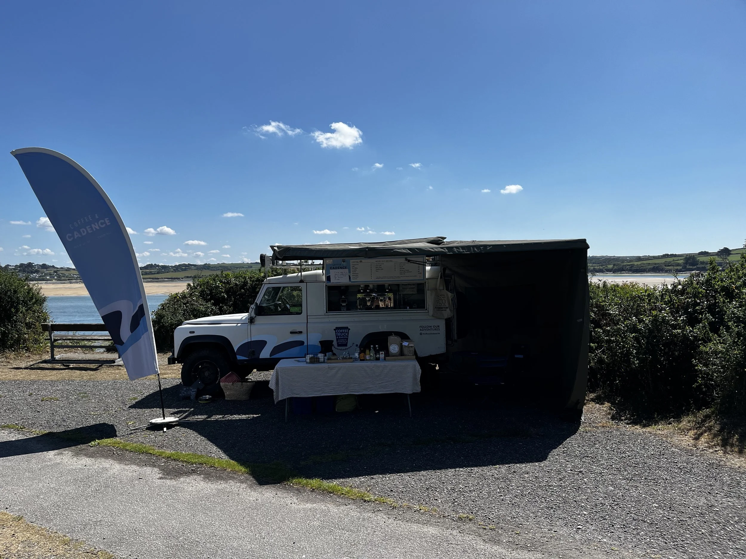 A mobile coffee stand set up next to a water body under a clear blue sky, with a flag displaying the 'Coffee & Cadence' logo and a table with coffee supplies.