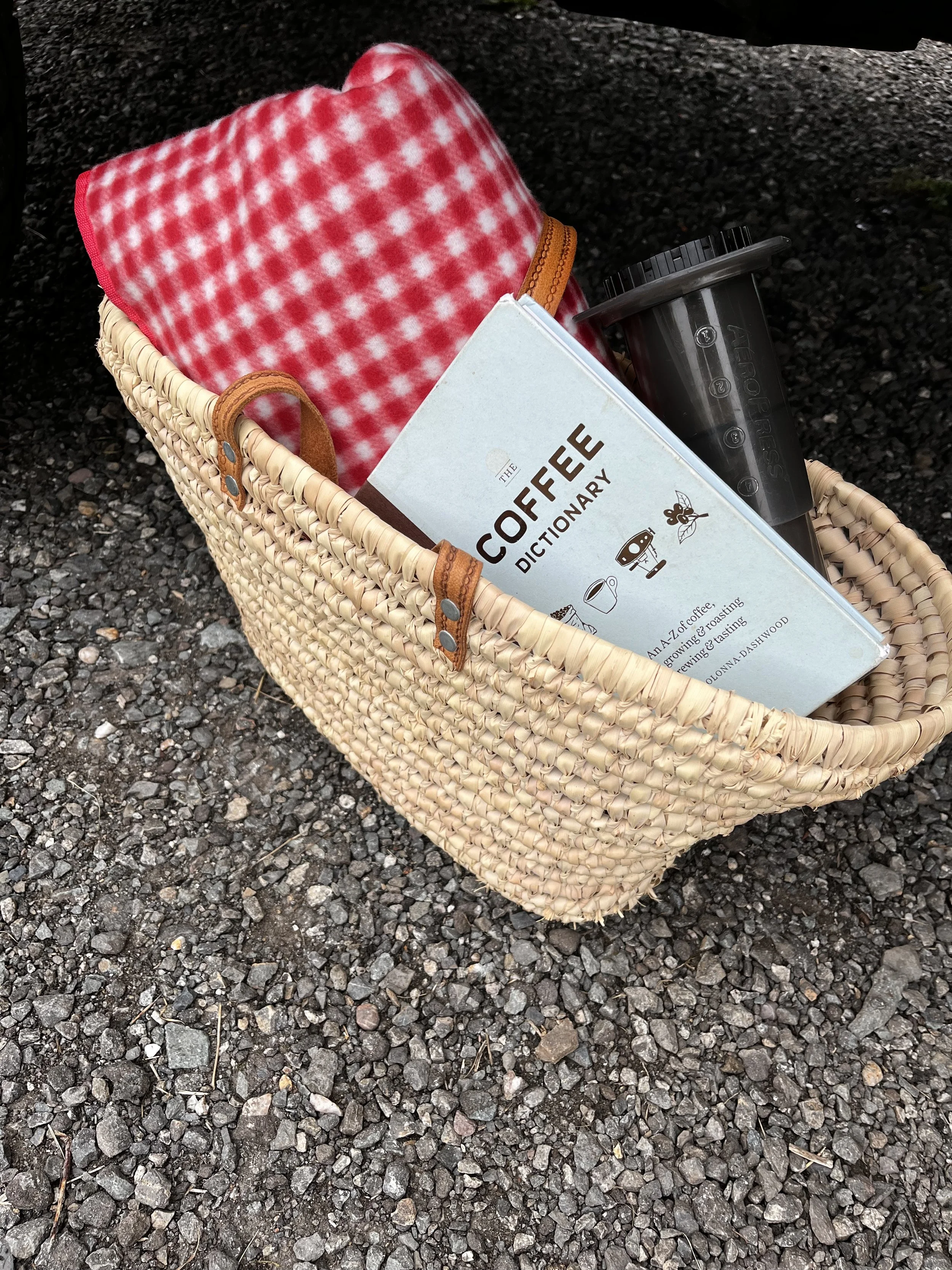 A woven straw basket containing a red and white checkered cloth, a book titled 'The Coffee Dictionary', a black reusable coffee cup, and a black travel tumbler, placed on a gravel surface.