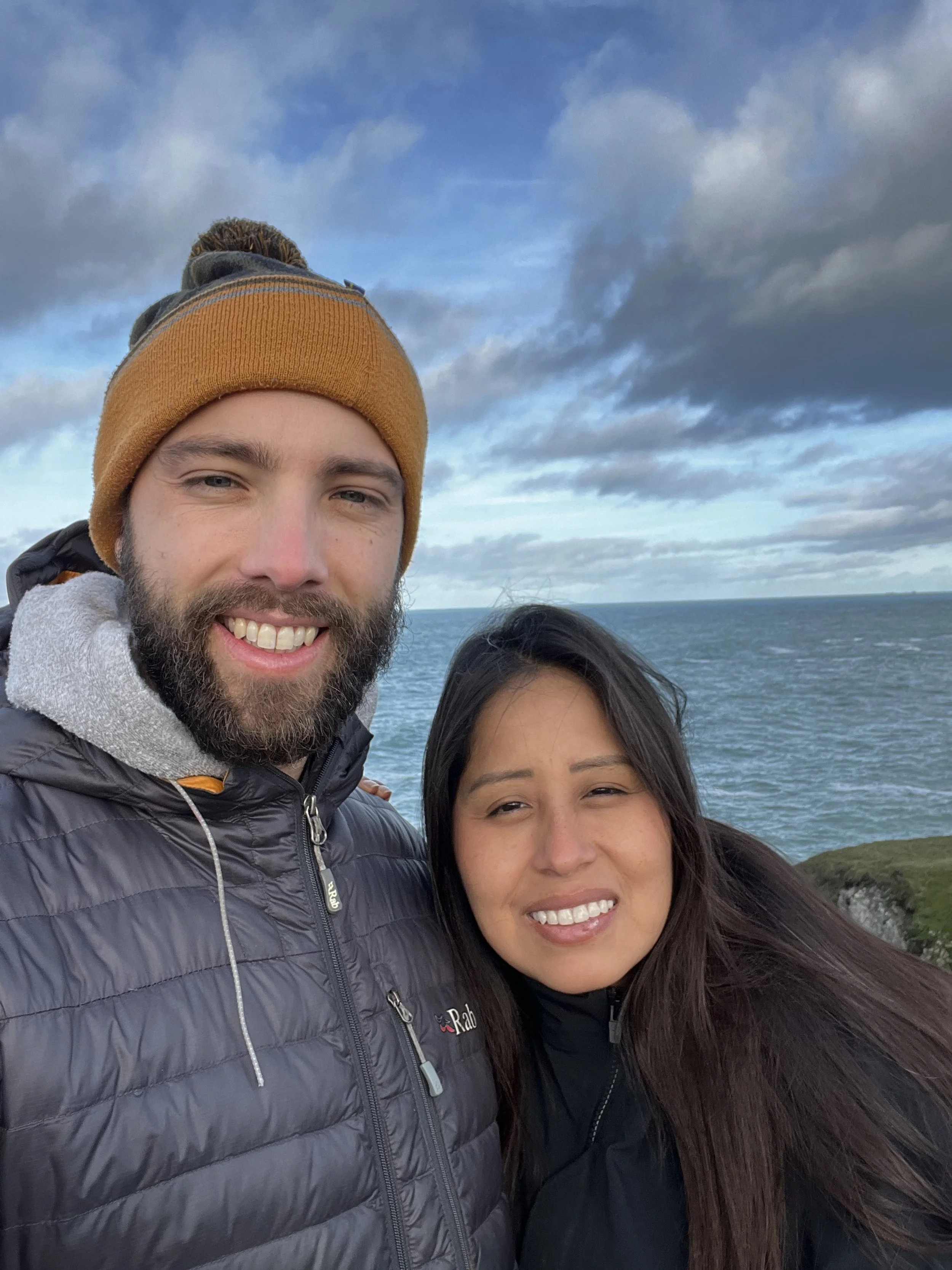 A smiling bearded man in a brown beanie and a black puffer jacket, standing next to a woman with long dark hair and a black jacket, with an ocean and cloudy sky in the background.