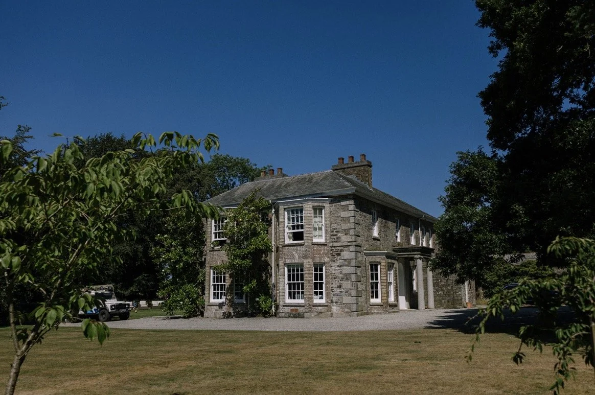 A large two-story house built with stone, surrounded by trees and grass, with a clear blue sky overhead.