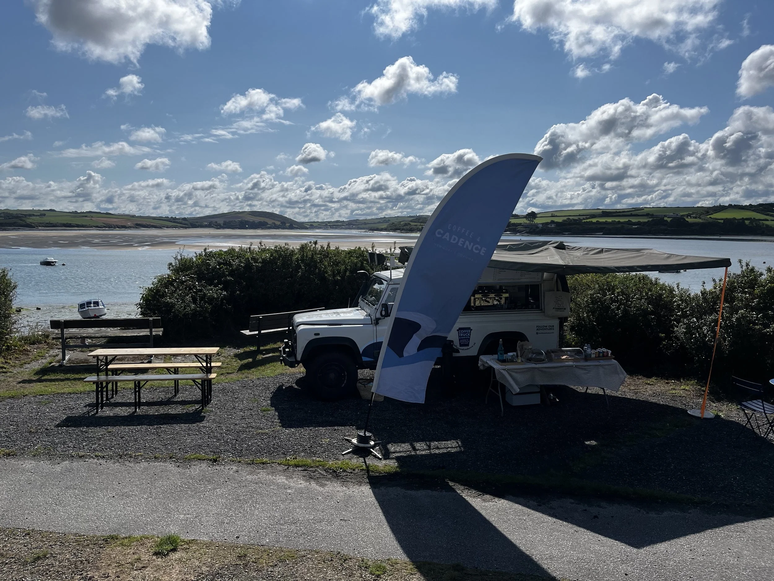A mobile coffee stand set up on a grassy area near a body of water, with benches and tables, and a sailboat and cars in the distance under a partly cloudy sky.