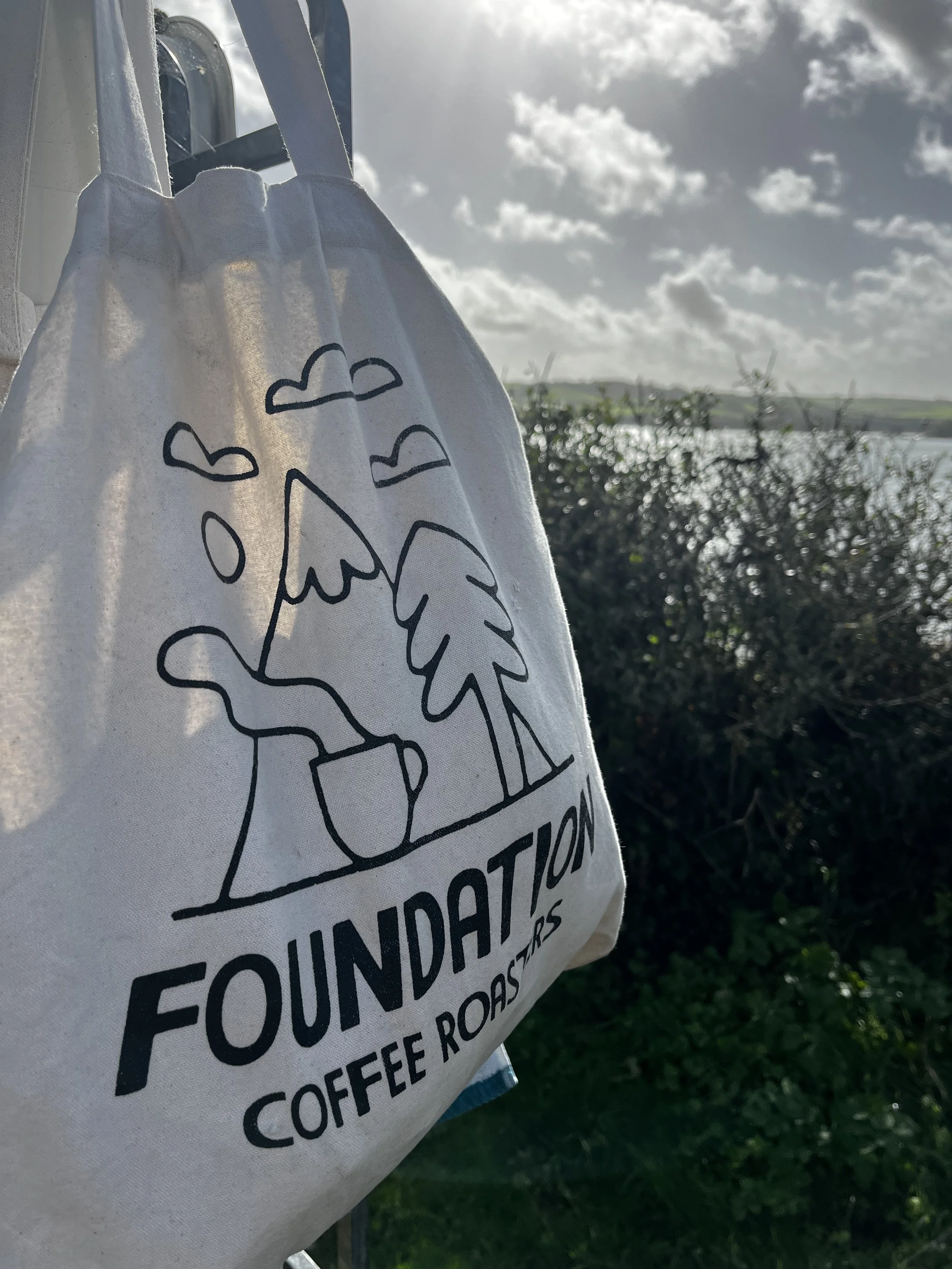 A white tote bag with black text and a simple graphic of a coffee cup and coffee beans, hanging outdoors against a cloudy sky and greenery background.