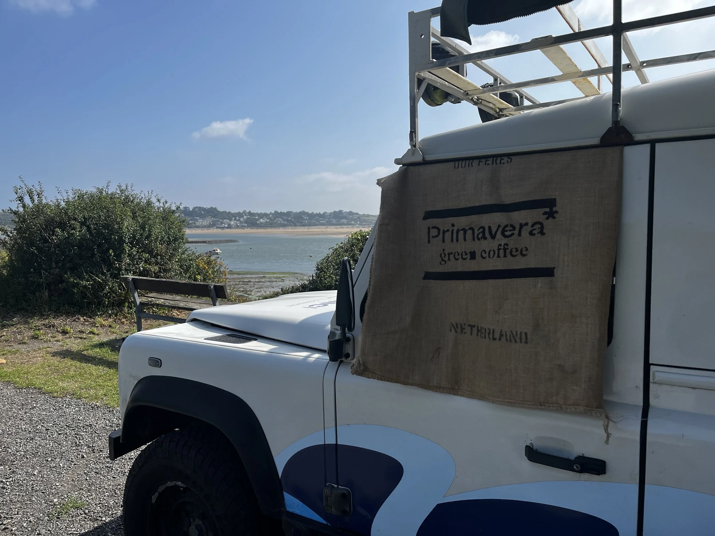 A white vehicle with a canvas banner reading 'Primavera green coffee Netherlands' parked near water with bushes, a bench, and a clear sky in the background.