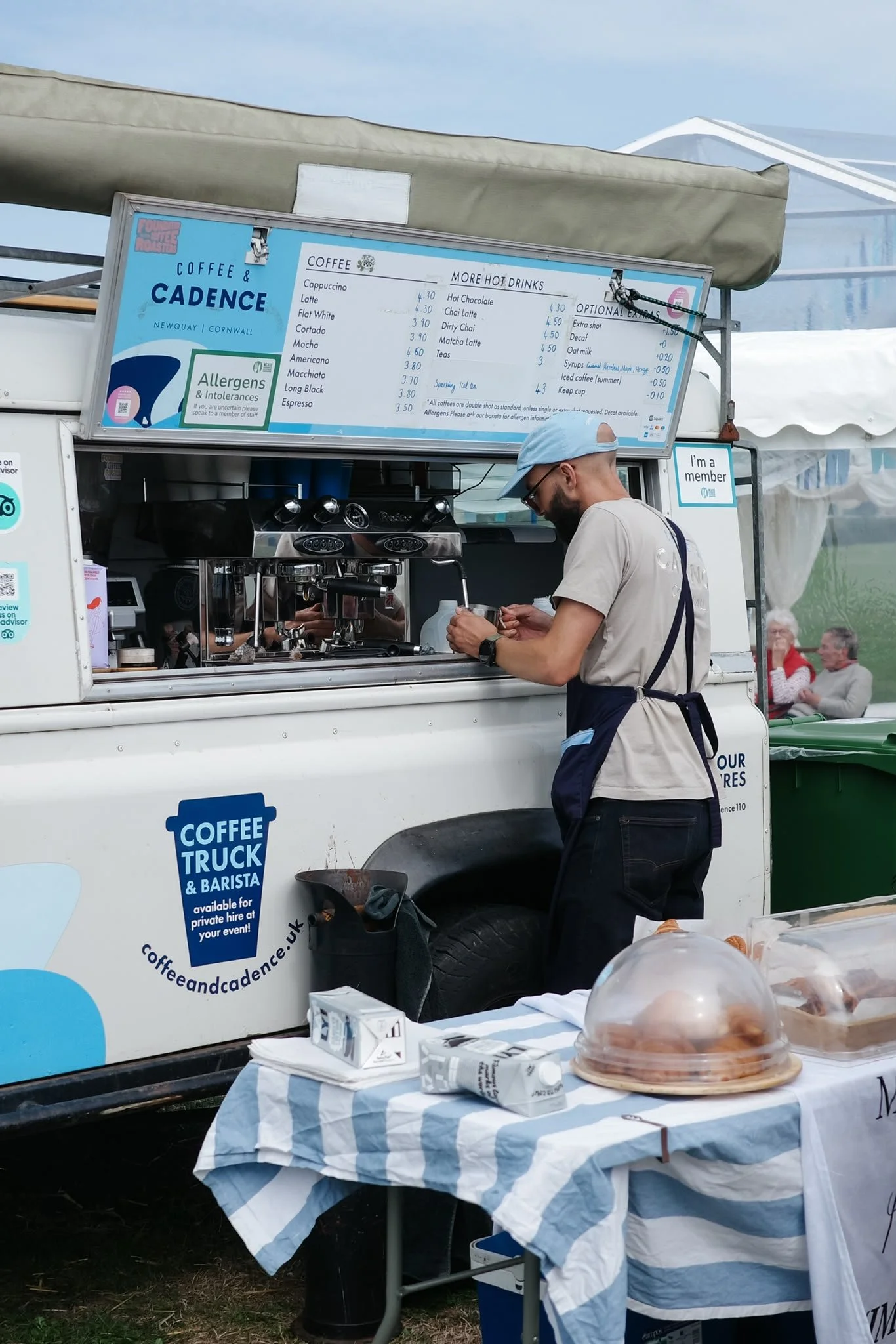 A man in a light blue cap and apron working at a coffee truck, with a menu board displaying various coffee and hot drinks, and a table with baked goods in front of him.