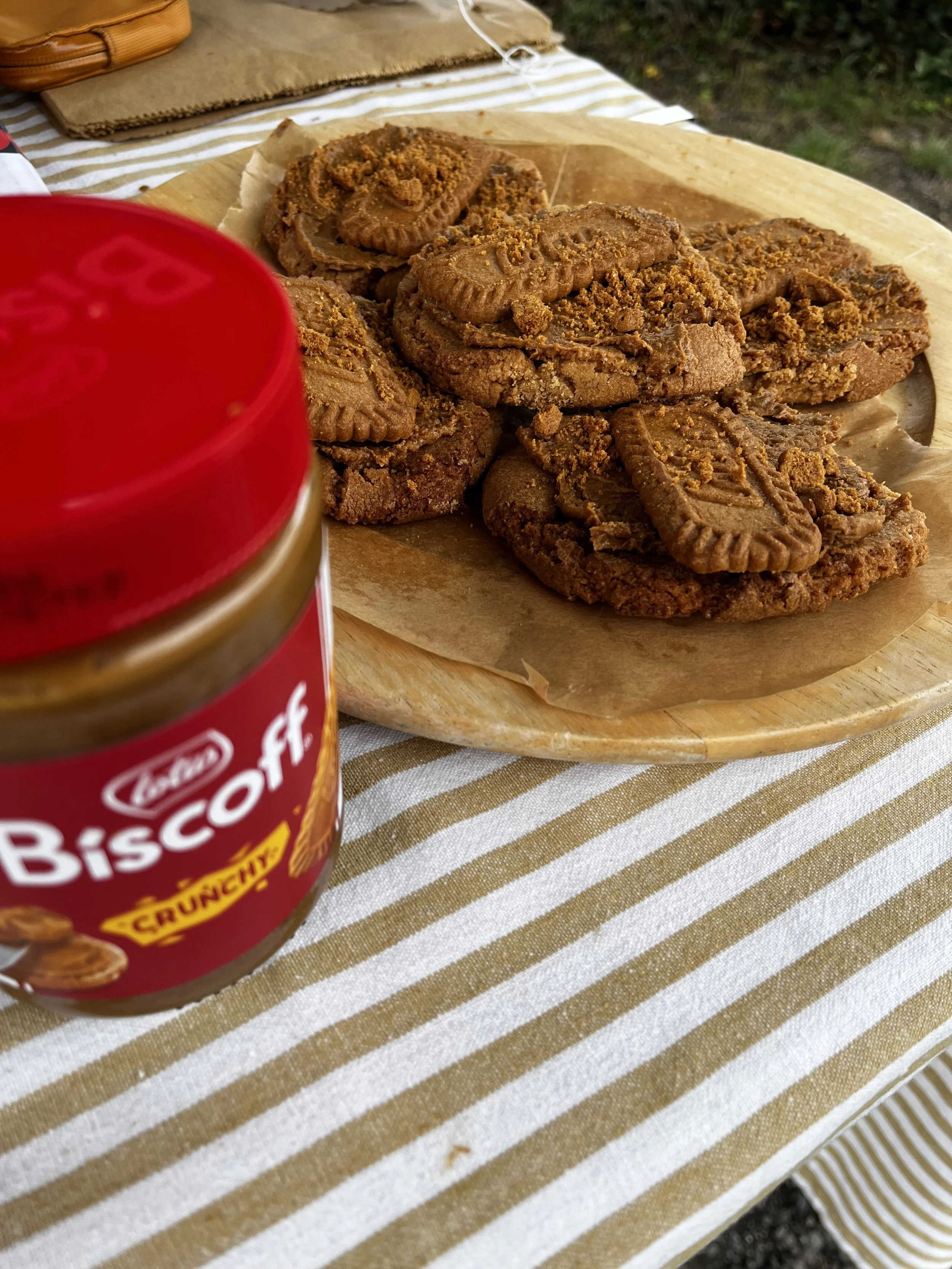 A wooden plate with peanut butter cookies on parchment paper, placed on a striped tablecloth, with a jar of peanut butter in the foreground.