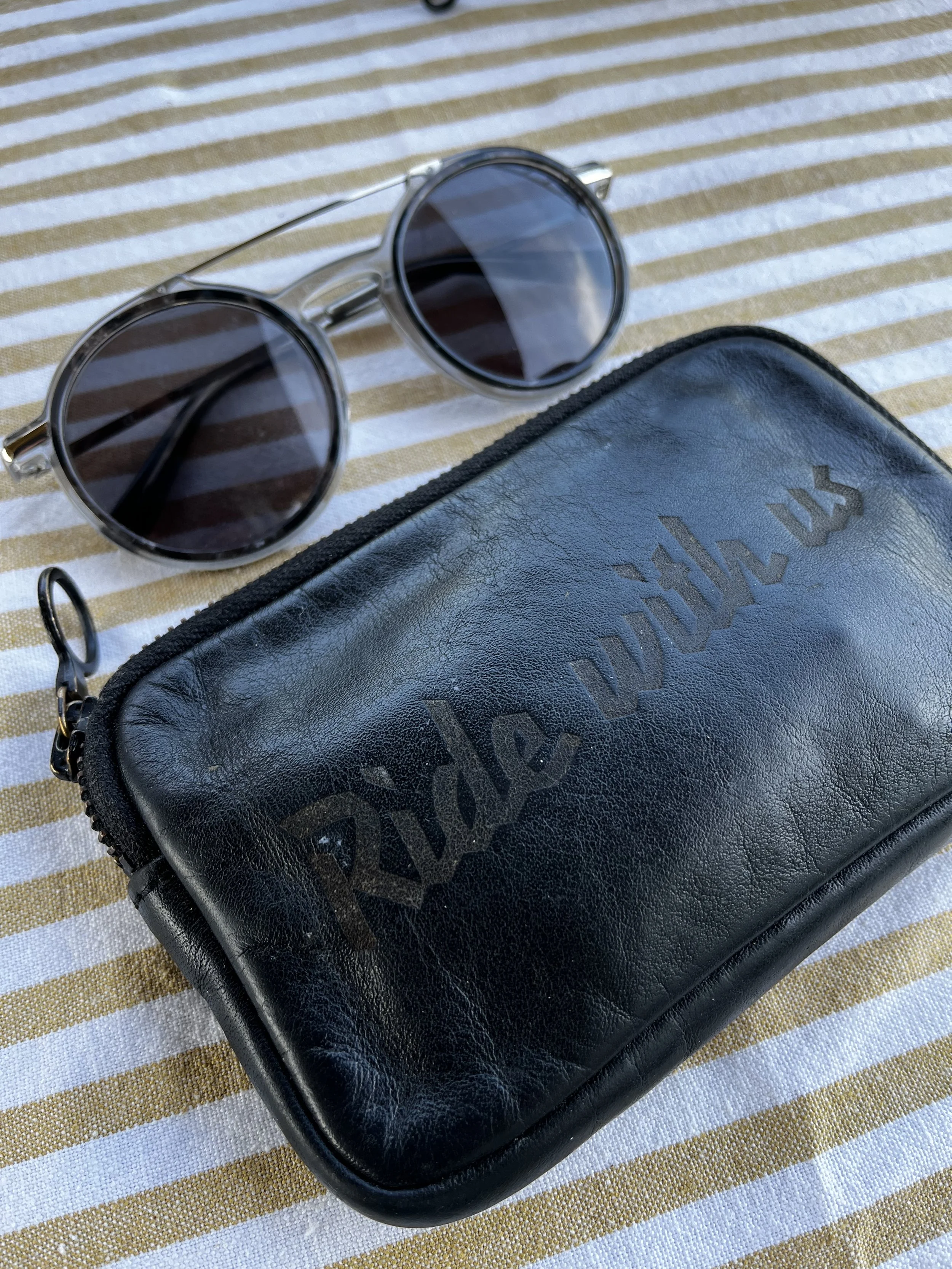 A pair of round sunglasses with dark lenses and silver frames resting on a striped fabric surface next to a black leather case with embossed text.