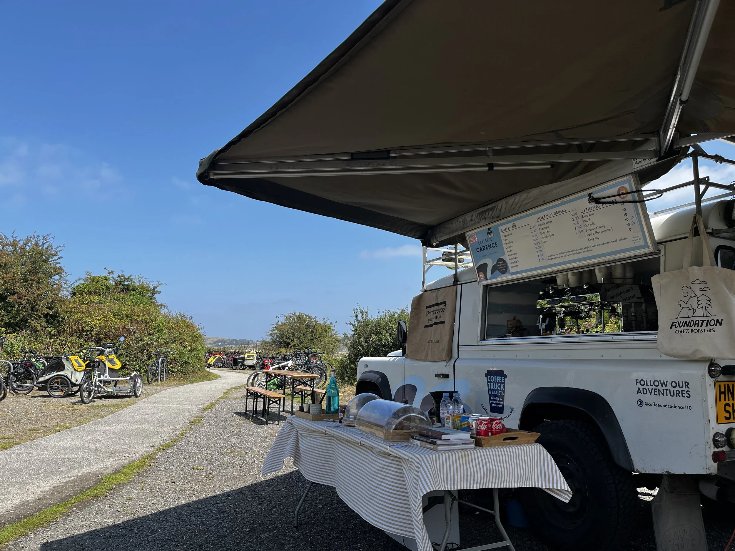 A white coffee truck set up outdoors with a striped tablecloth on a table, in front of a scenic view with bicycles parked along a path and lush green bushes, under a clear blue sky.