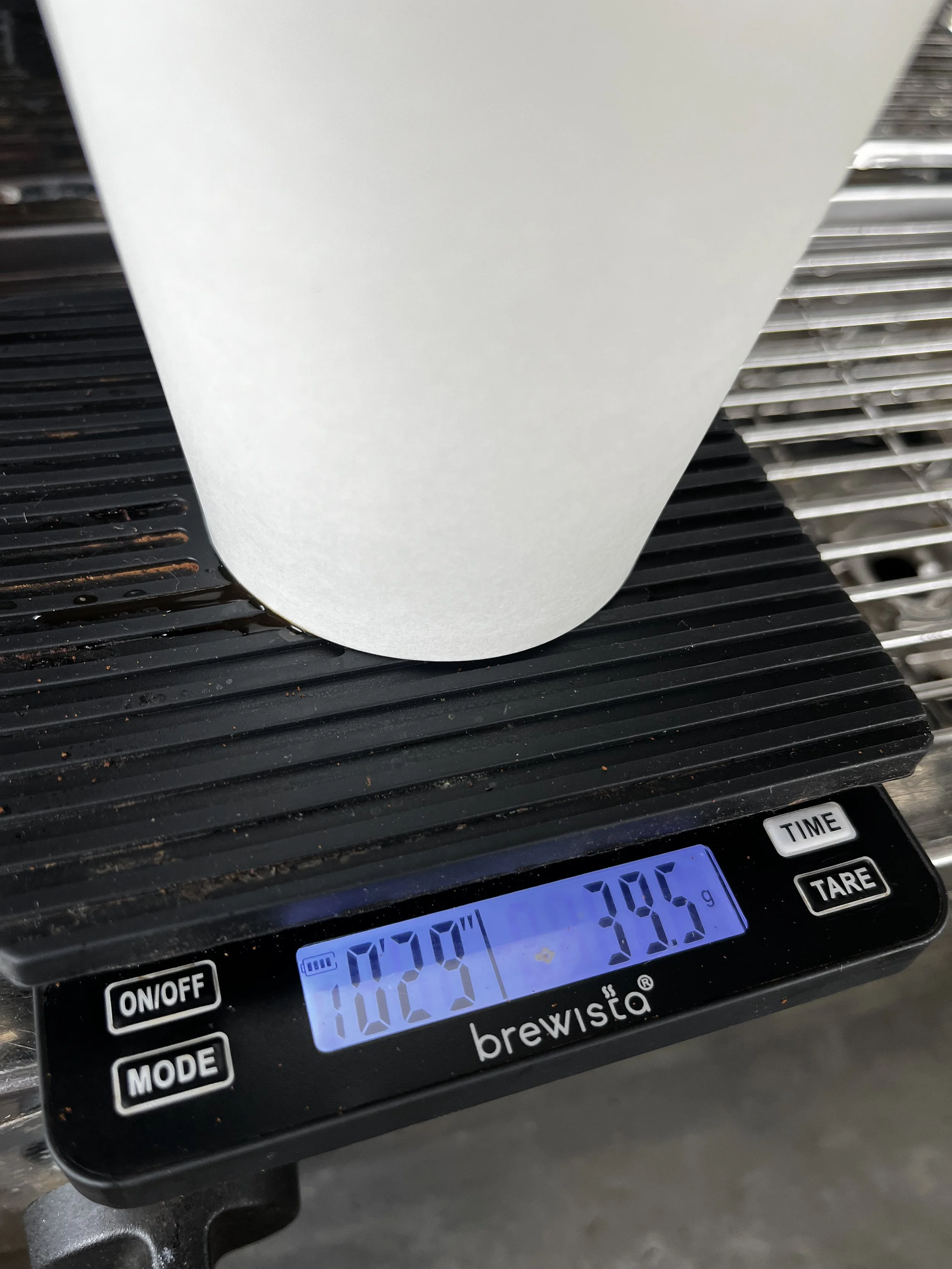 A digital kitchen scale displaying a weight of 38.5 grams, with a white container on top, placed on a black surface with ridges.