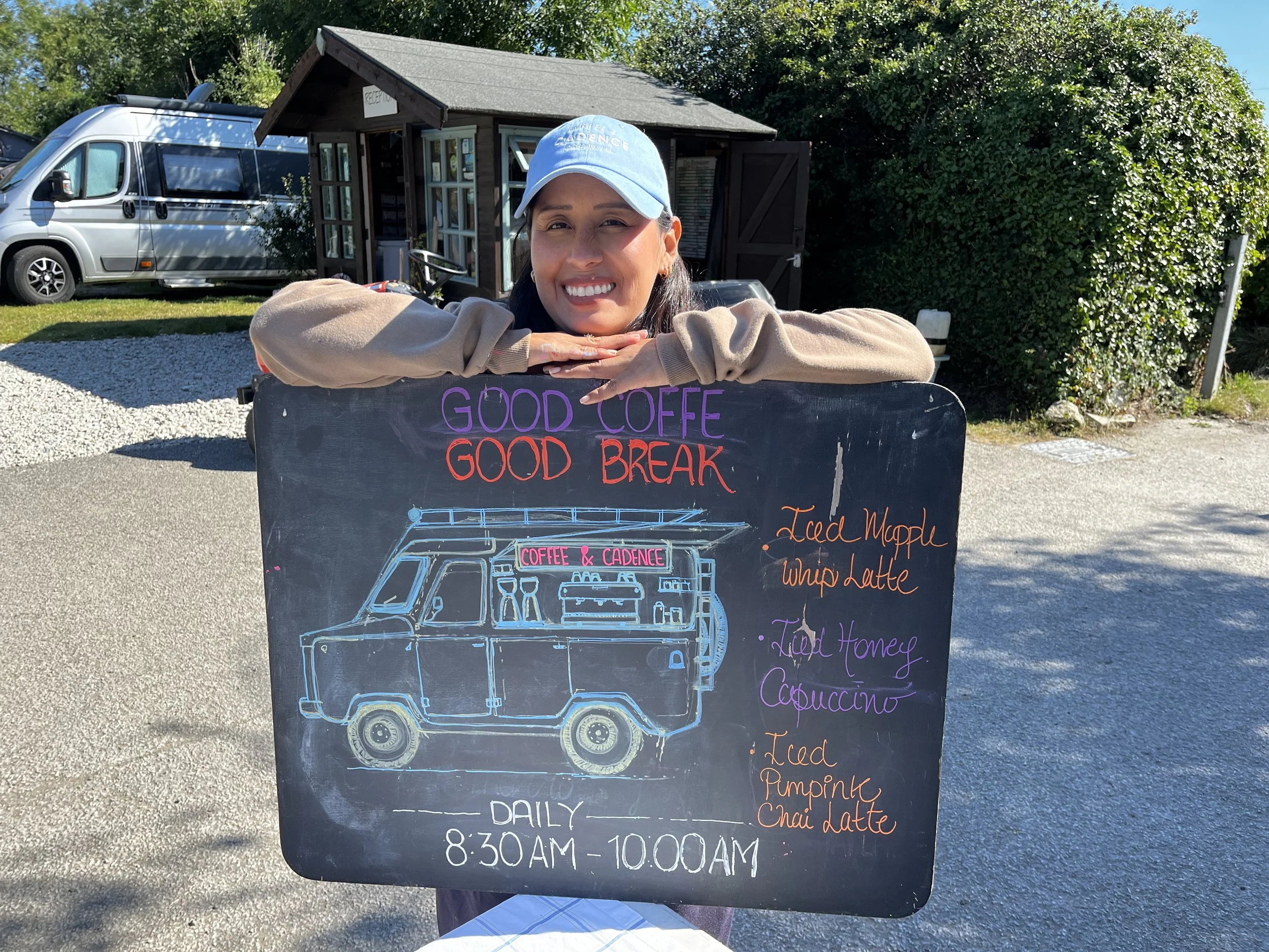 A woman with dark hair wearing a light blue baseball cap and beige jacket, smiling with her arms resting on a blackboard sign. The sign has colorful drawings of a food truck and lists menu items like iced maple, whipped latte, and chai latte. She is 