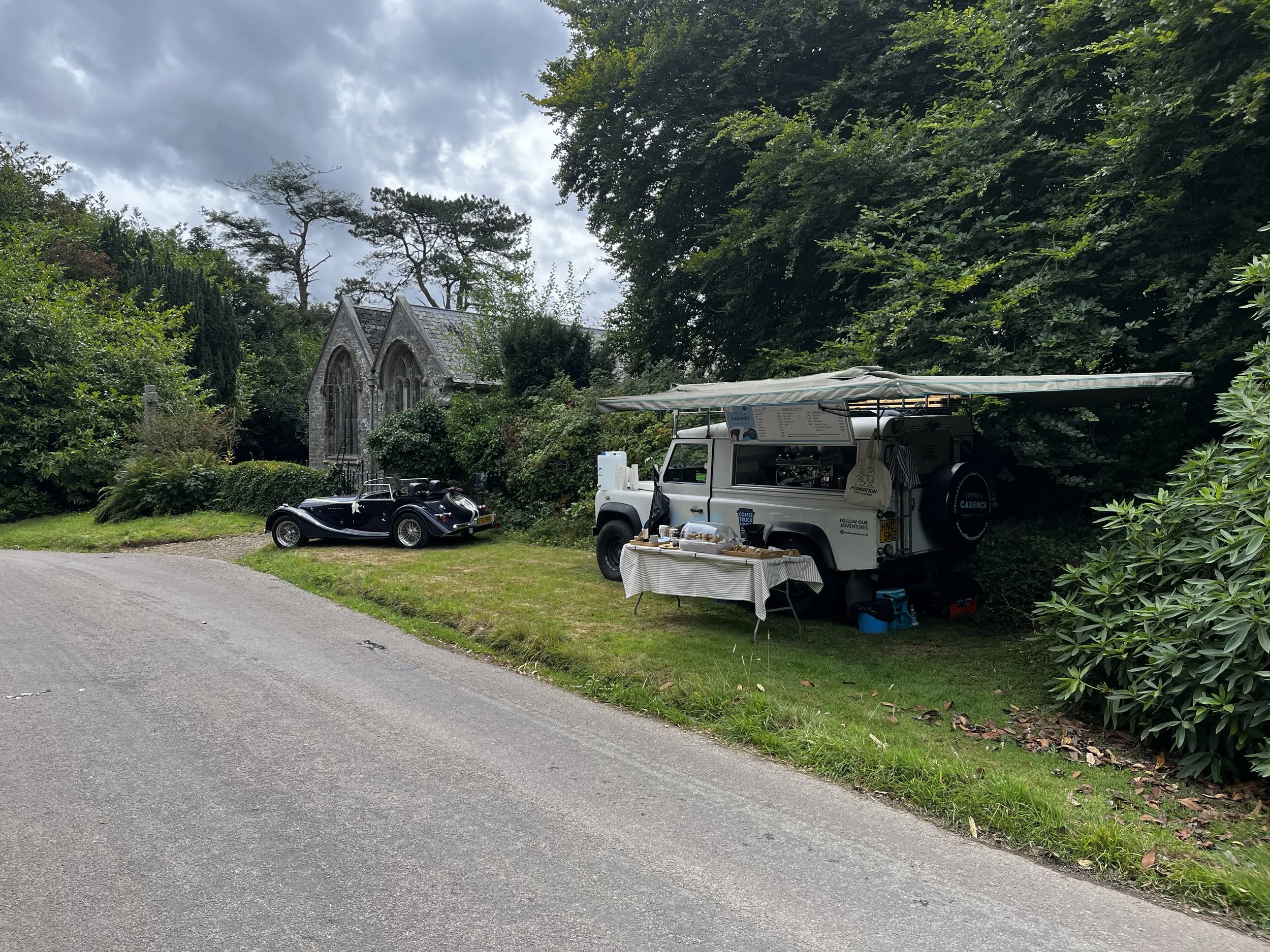 A vintage black car parked near a small church, with a mobile coffee stand set up under a canopy nearby, in a lush green area with cloudy sky overhead.