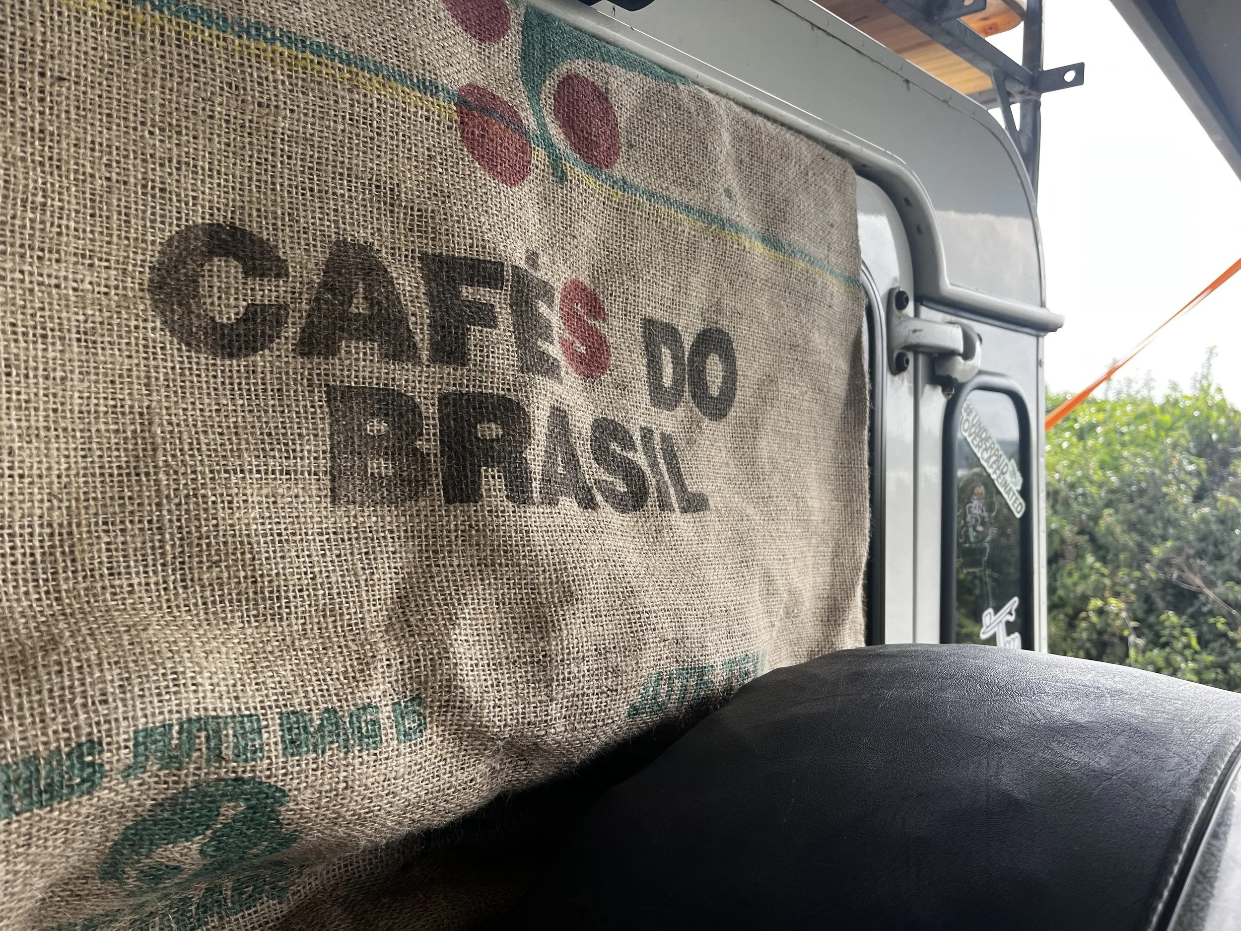 Close-up view of a burlap coffee bag with the words 'Cafés do Brasil' printed on it, inside a vehicle with a black seat and some greenery visible outside the window.
