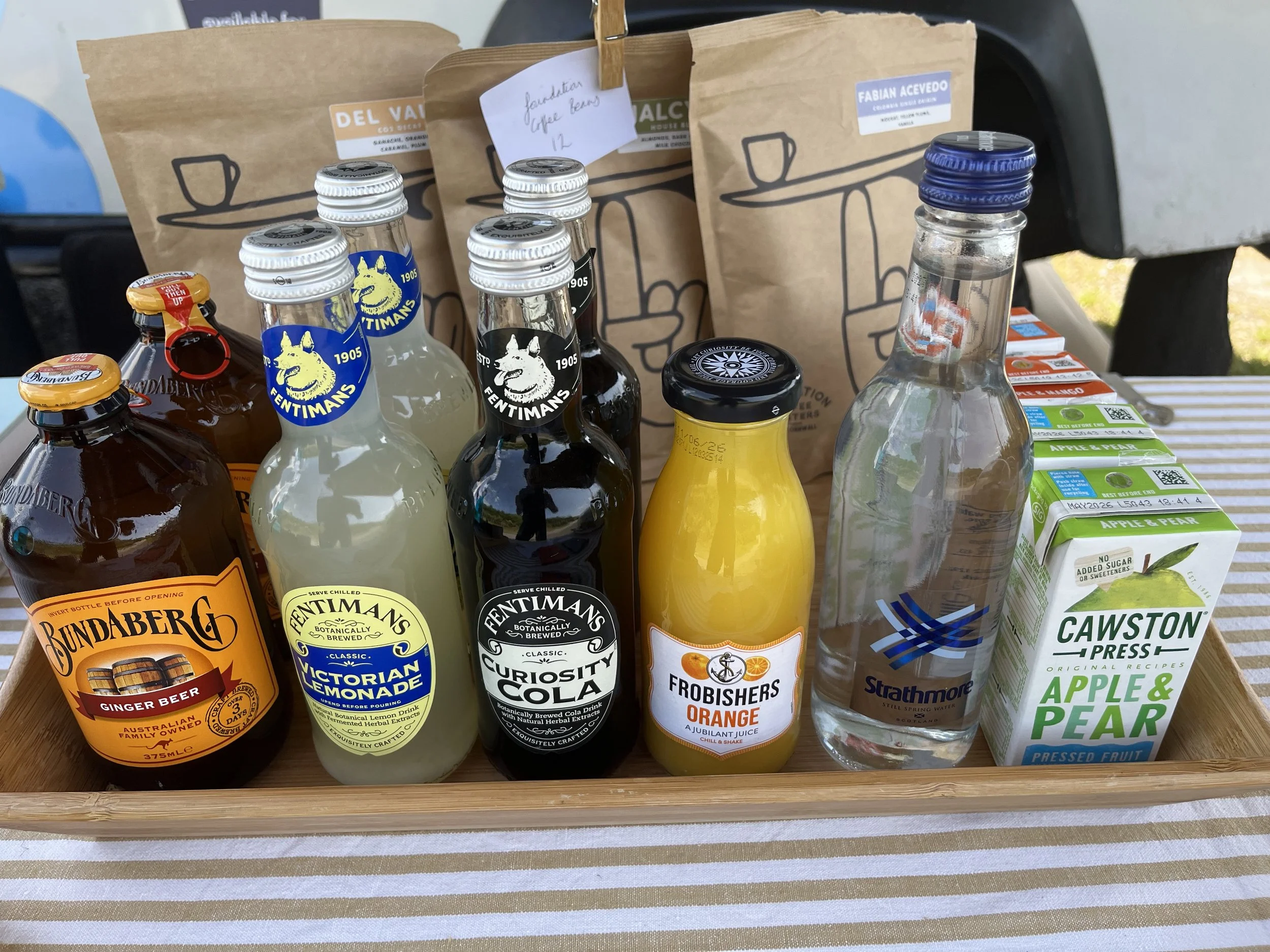 Assorted beverages including ginger beer, Victoria lemonade, curiosity colada, orange juice, water, and apple and pear pressed fruit juice in a wooden tray on a striped tablecloth.