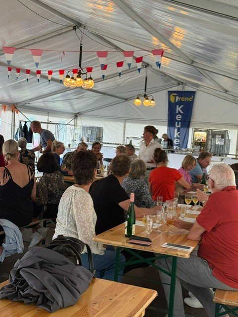 People gathering inside a large tent for a social event, sitting at long tables, with bunting and lights hanging from the ceiling, and a bar area in the background.
