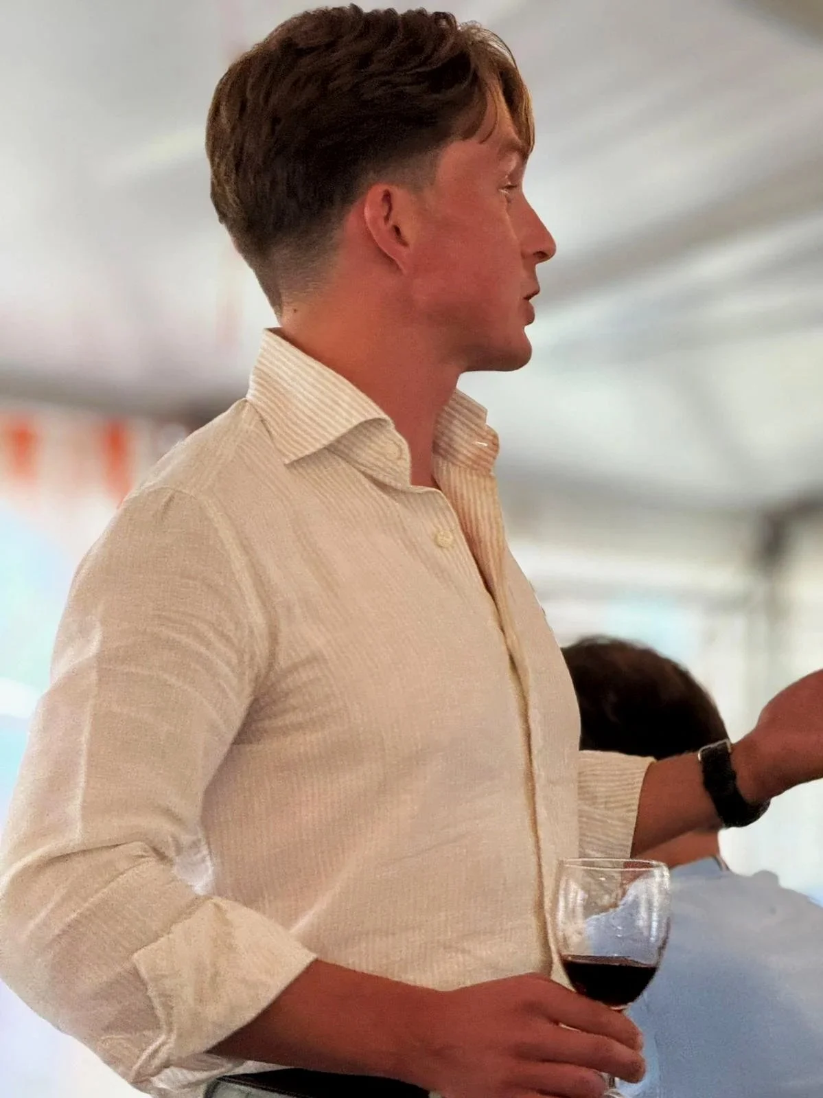 A young man with brown hair and fair skin is holding a glass of red wine while standing indoors, wearing a light-colored, long-sleeved, striped button-up shirt.