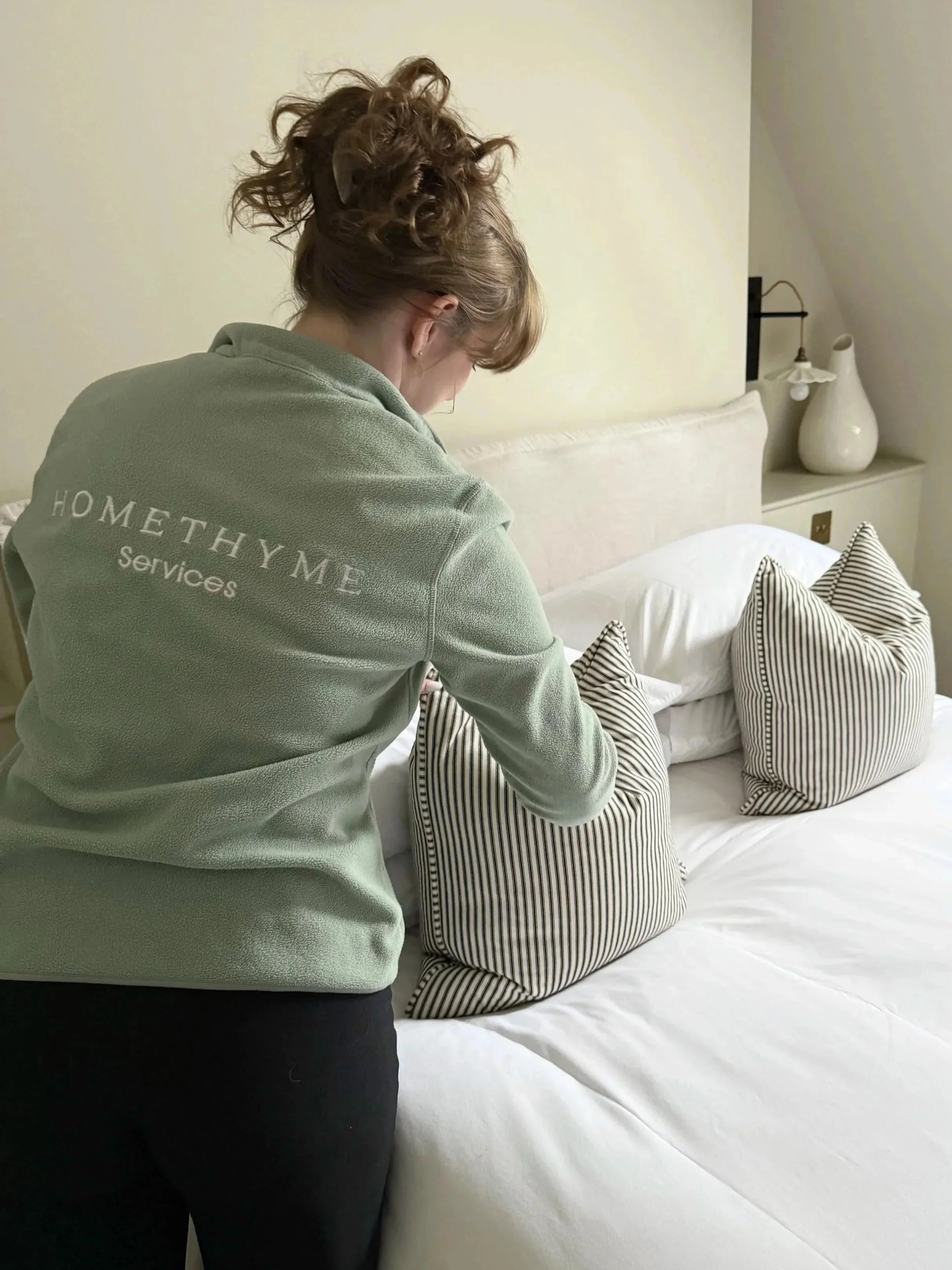 A woman with curly hair wearing a green fleece jacket with 'HOMETHYME Services' on the back, making a bed with striped pillows in a neutral-toned bedroom.