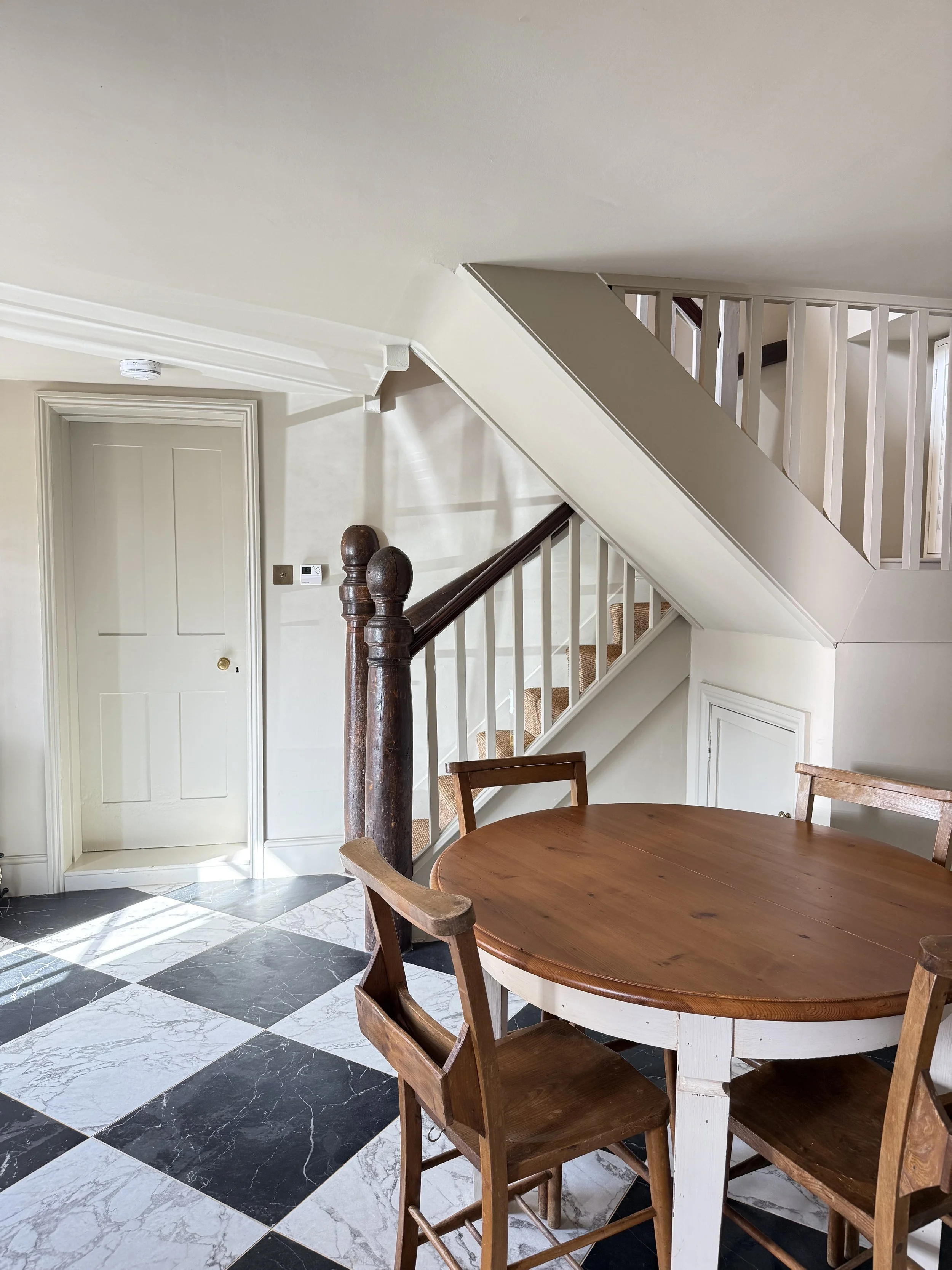 A dining area next to a staircase with a round wooden table and four chairs, marble-tiled floor, and a white door.