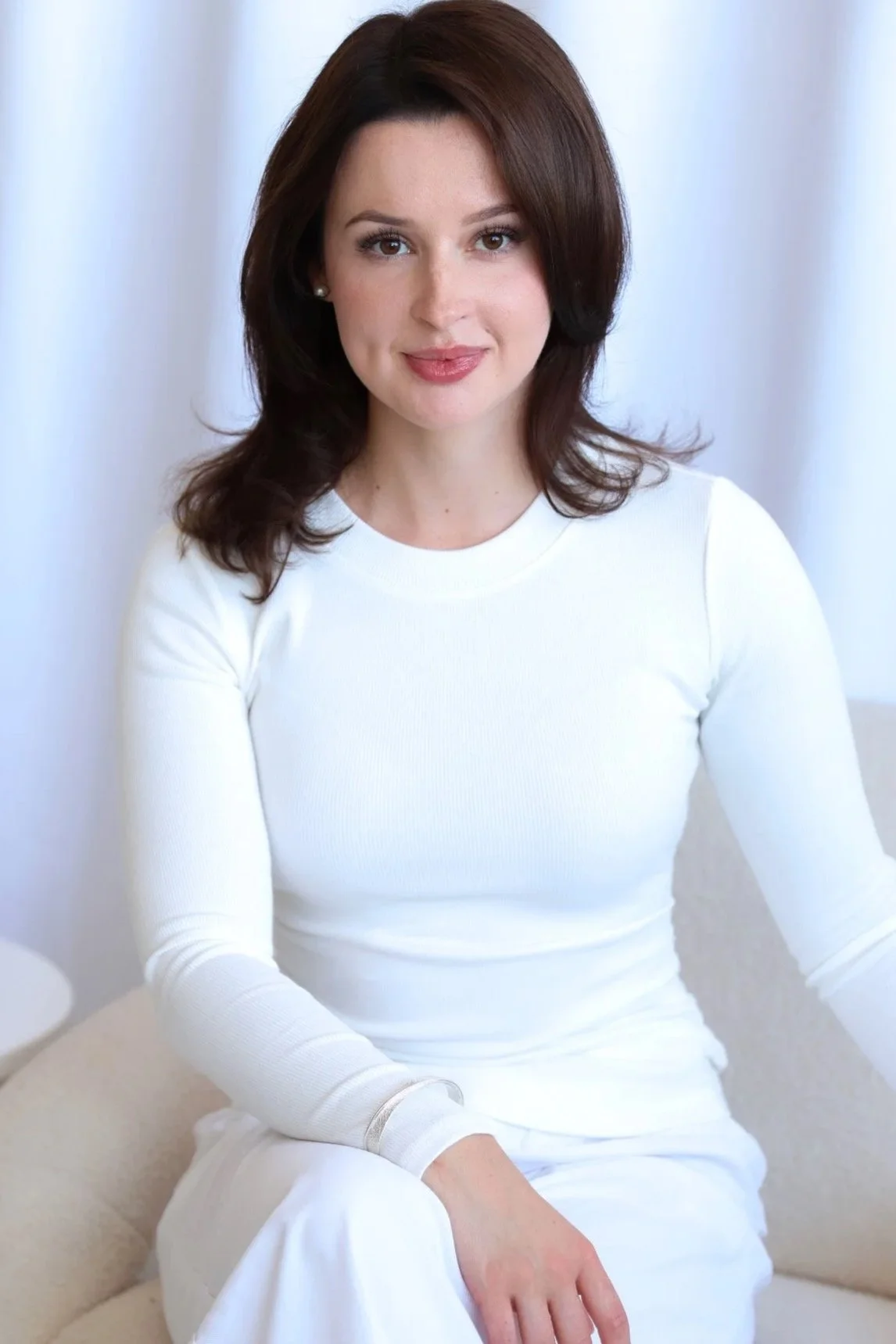 A woman with shoulder-length brown hair, fair skin, wearing a white long-sleeve top, sitting on a beige sofa in front of a light-colored curtain.