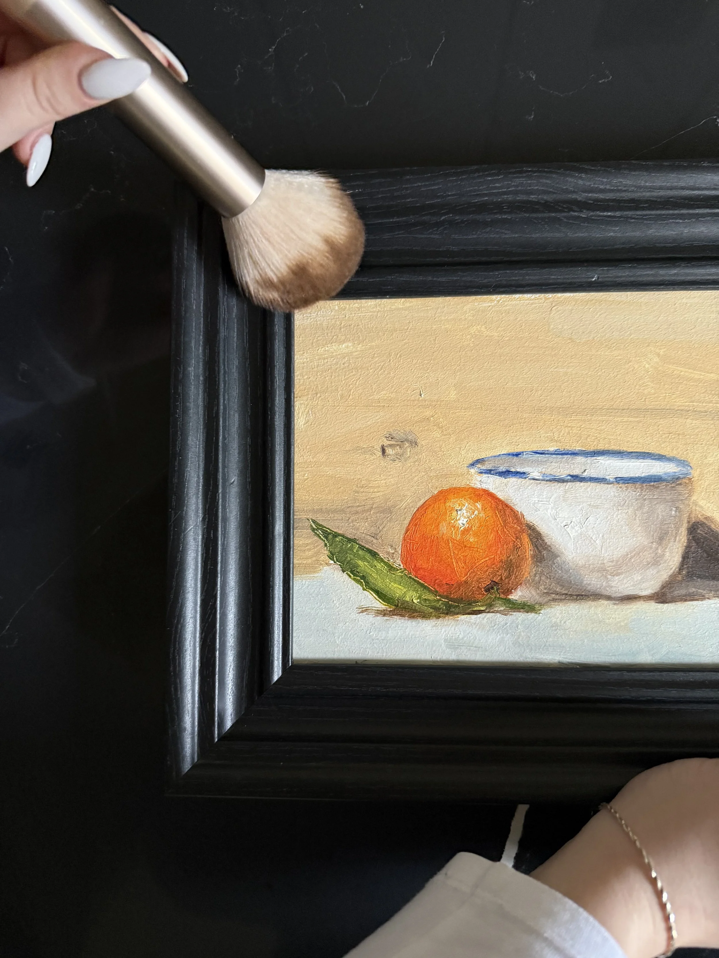 A person is brushing a painting of a still life with an orange, a green leaf, and a white bowl with a blue rim.