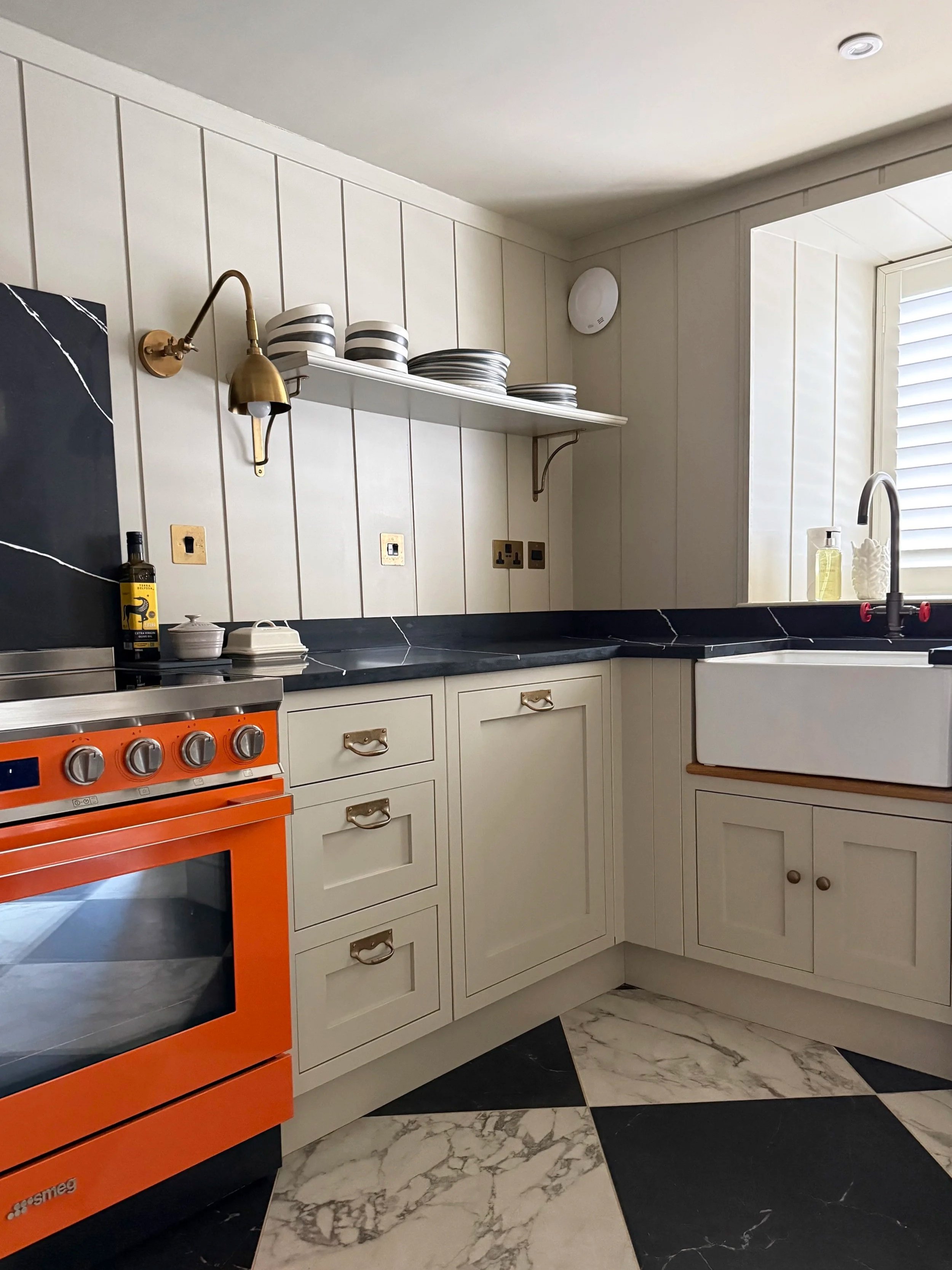 Kitchen with white cabinets, black marble countertop, orange oven, open shelving with black and white dishes, marble floor tiles, farmhouse sink, and window with shutters.