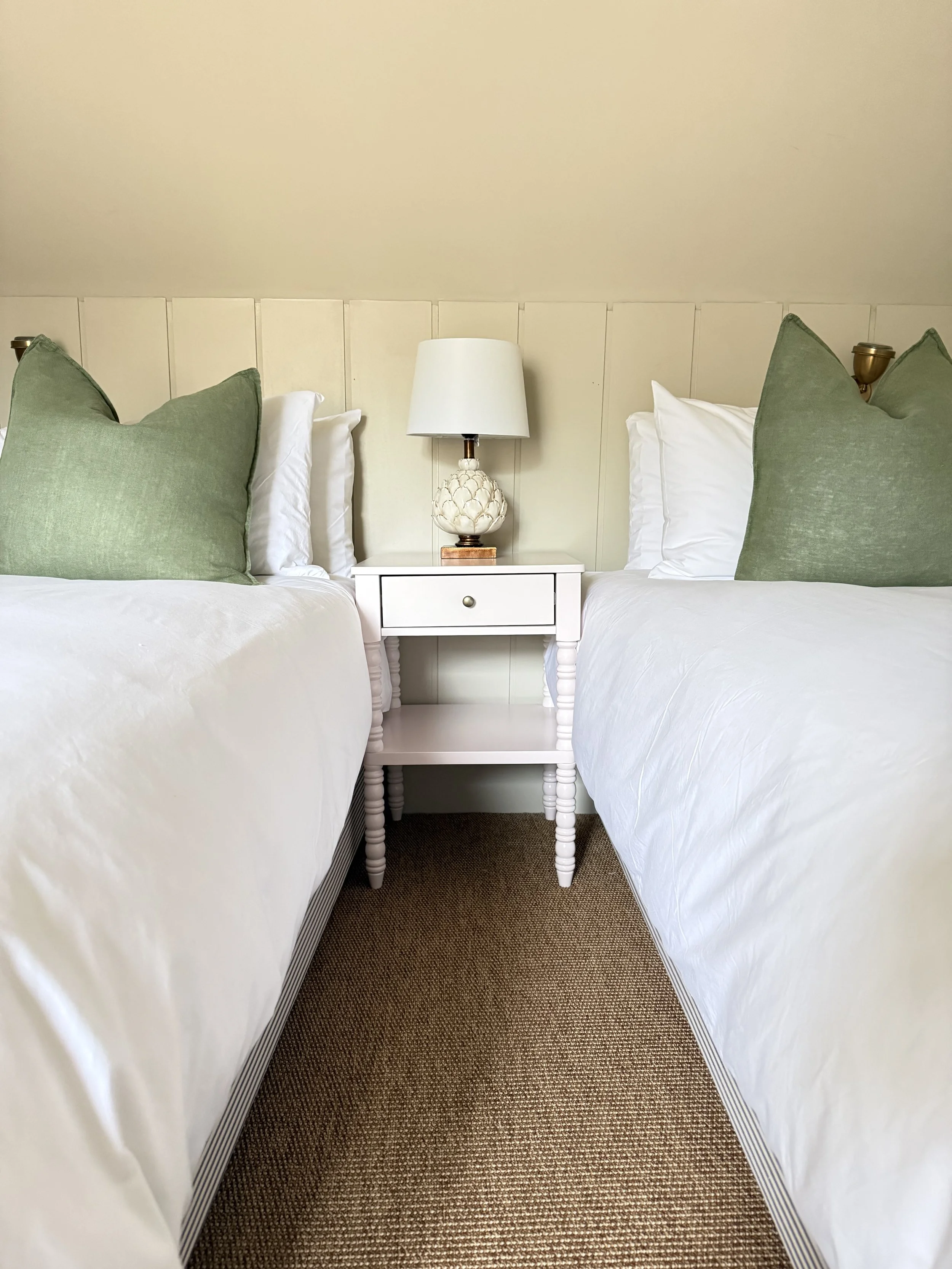 Two beds with white bedding and green pillows separated by a white nightstand with a white lamp on top in a bedroom.