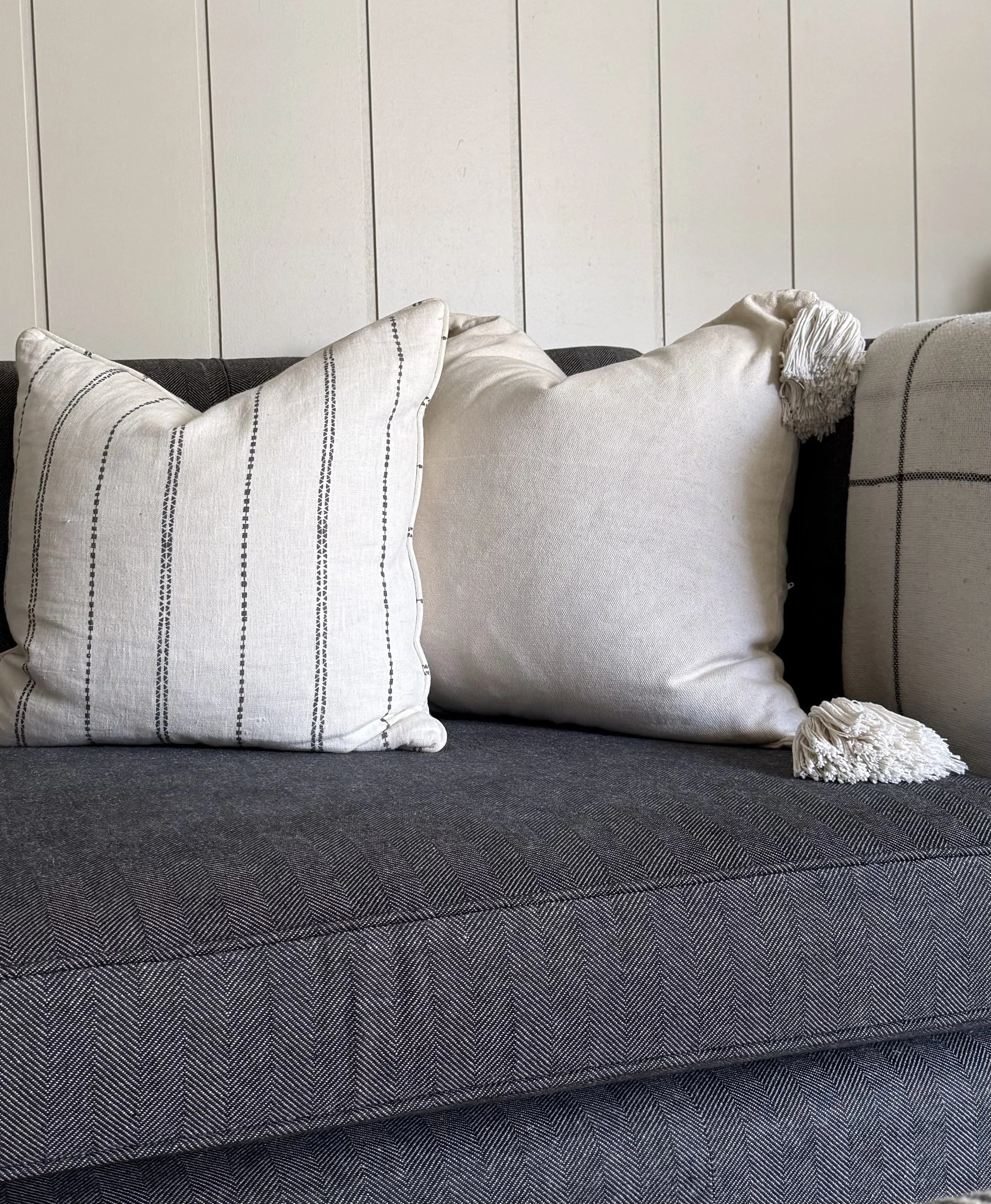 Close-up of a dark gray upholstered sofa with three cream-colored pillows, one with tassels, against a white wooden wall.
