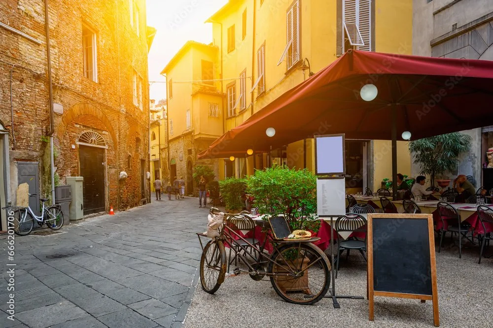 Outdoor restaurant seating with tables, chairs, and a large red umbrella on a narrow street in a European town, with buildings, pedestrians, and bicycles.