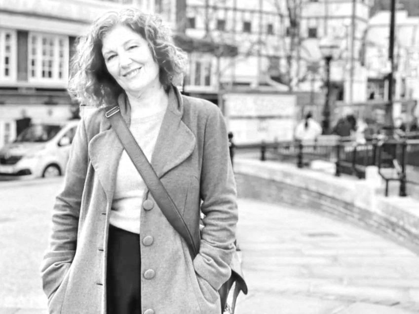 Black and white photo of a smiling woman with curly hair standing outdoors near a city street with cars and buildings in the background.