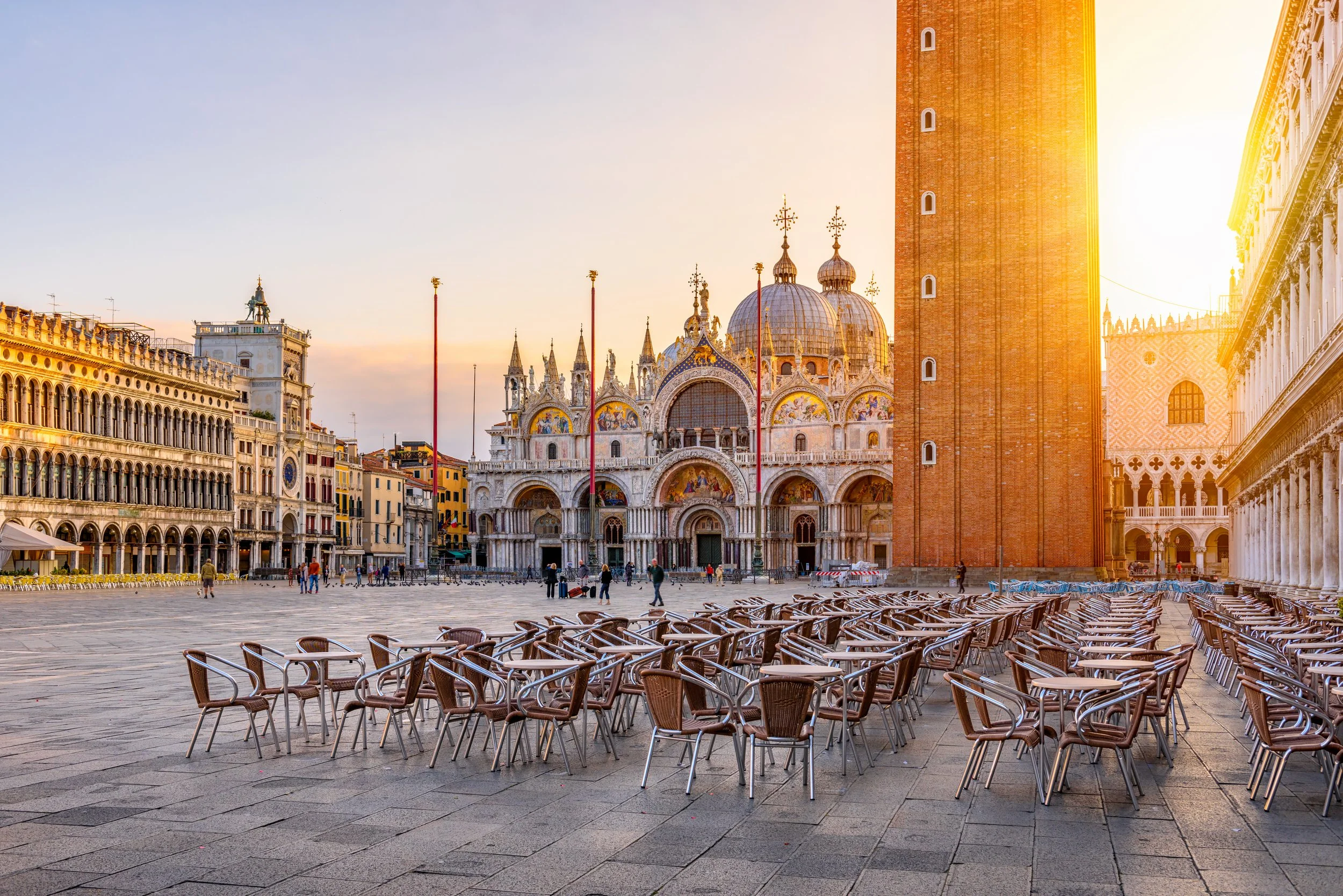 Empty outdoor plaza in Venice, Italy, with numerous chairs arranged in rows, historic buildings, and the Basilica di San Marco with its domes and intricate architecture in the background, bathed in warm sunlight.