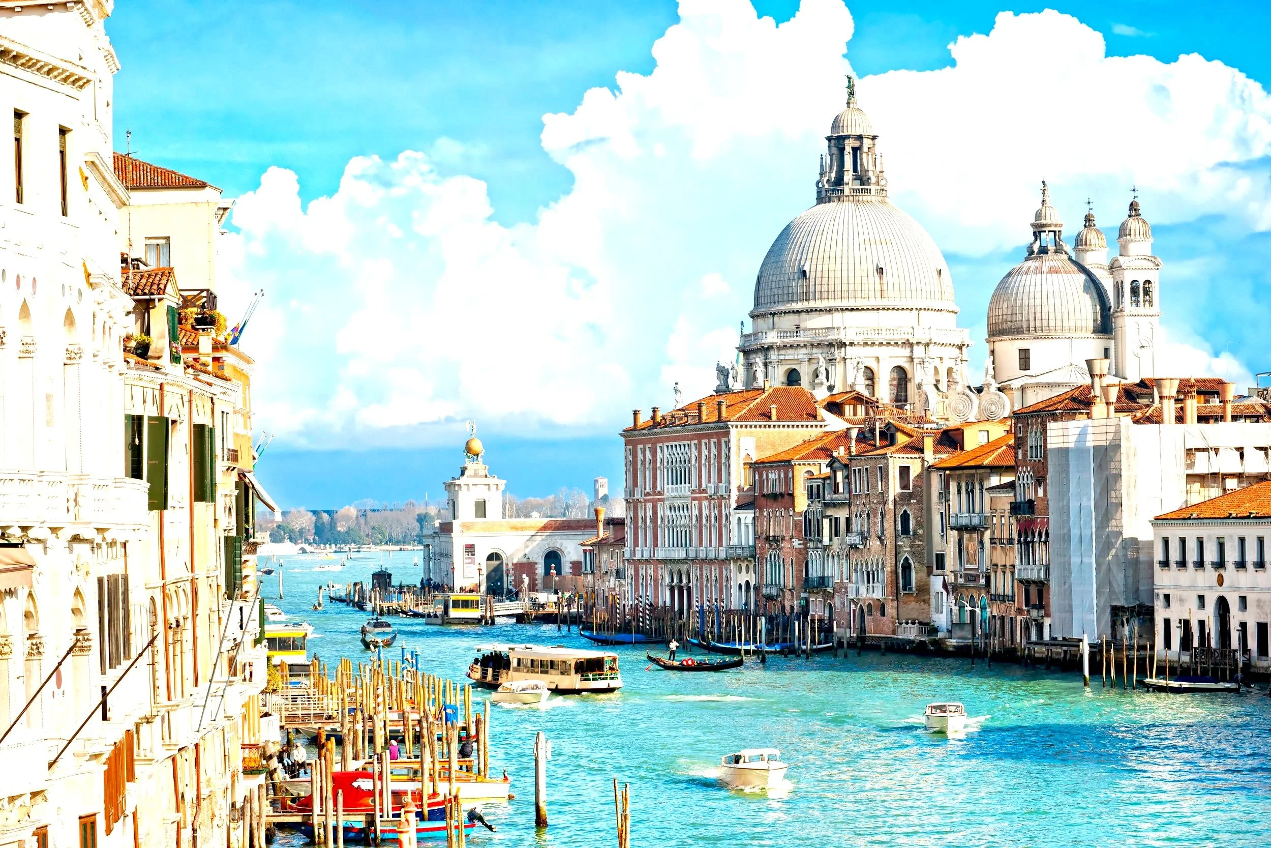 View of the Grand Canal in Venice, Italy with historic buildings, boats, and the Basilica di Santa Maria della Salute in the background under a partly cloudy sky.