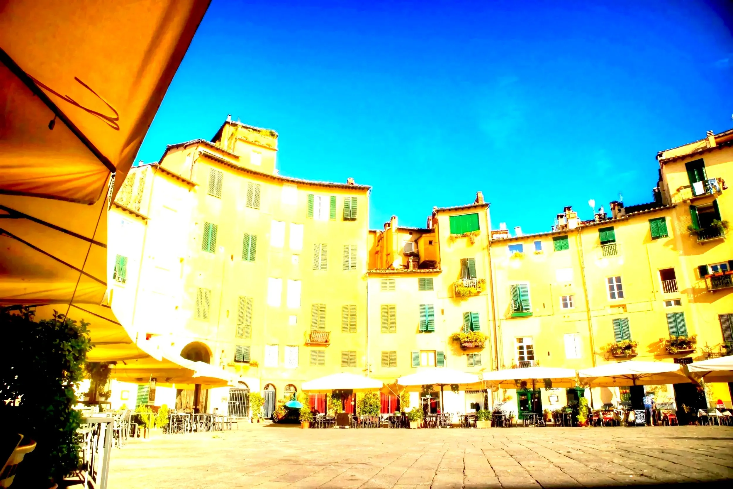 Yellow buildings with green shutters and flowers in window boxes, outdoor café with tables and umbrellas, blue sky, and a stone-paved square.
