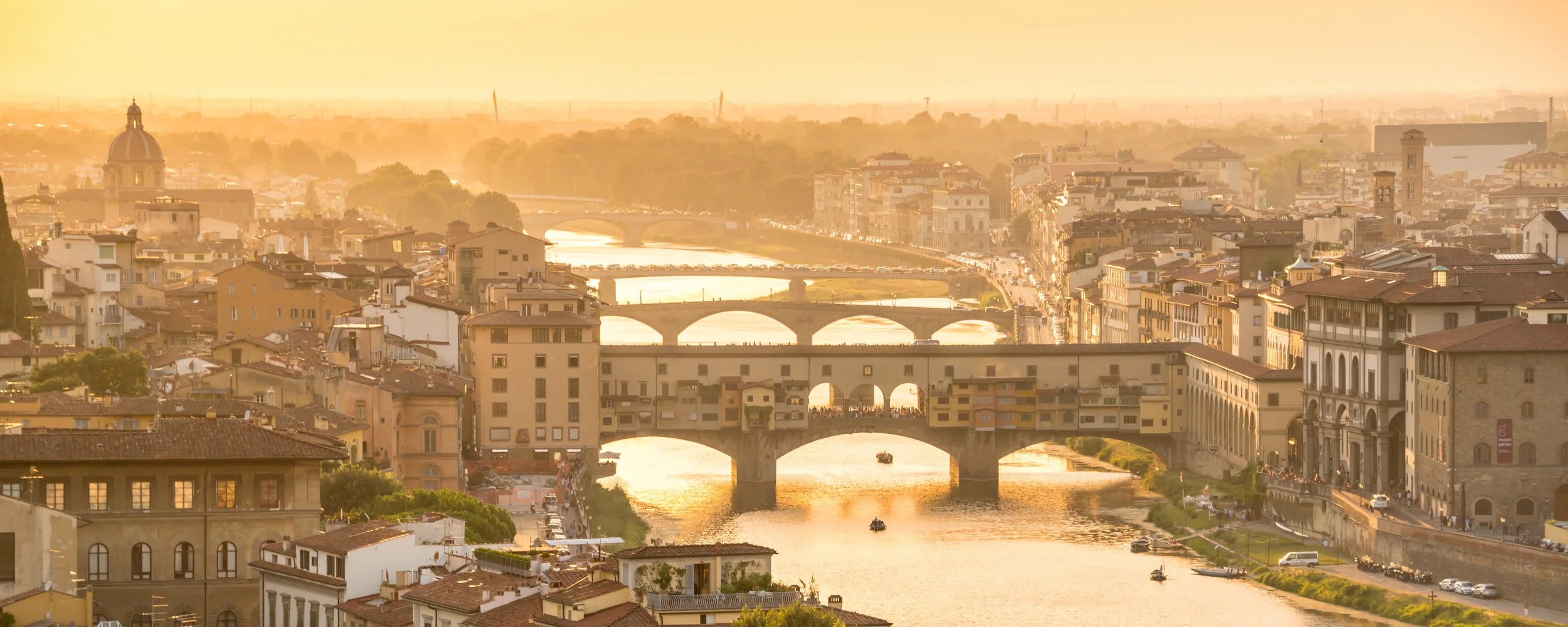 Sunset view of Florence, Italy, with historic bridges crossing the Arno River and city buildings in warm, golden light.